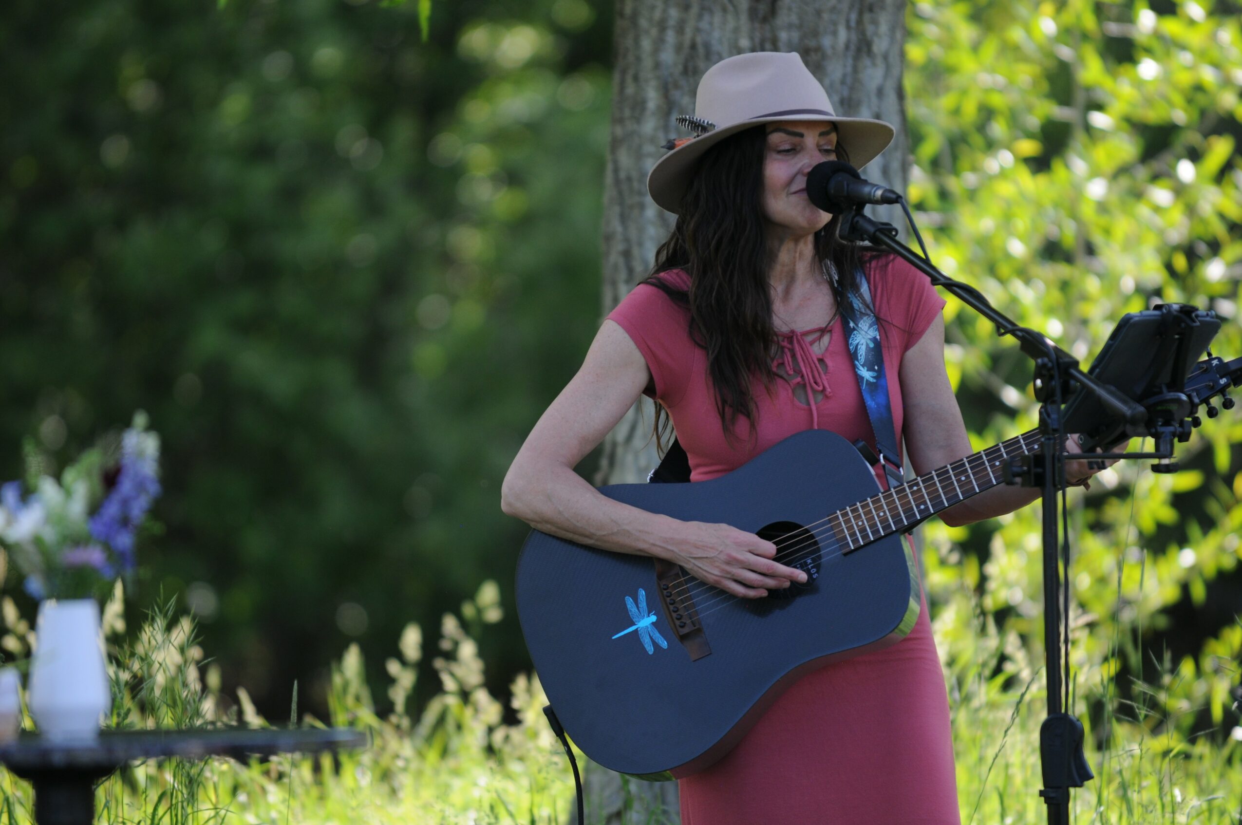 Woman wearing a hat and sunglasses playing guitar and singing outdoors with greenery in background.