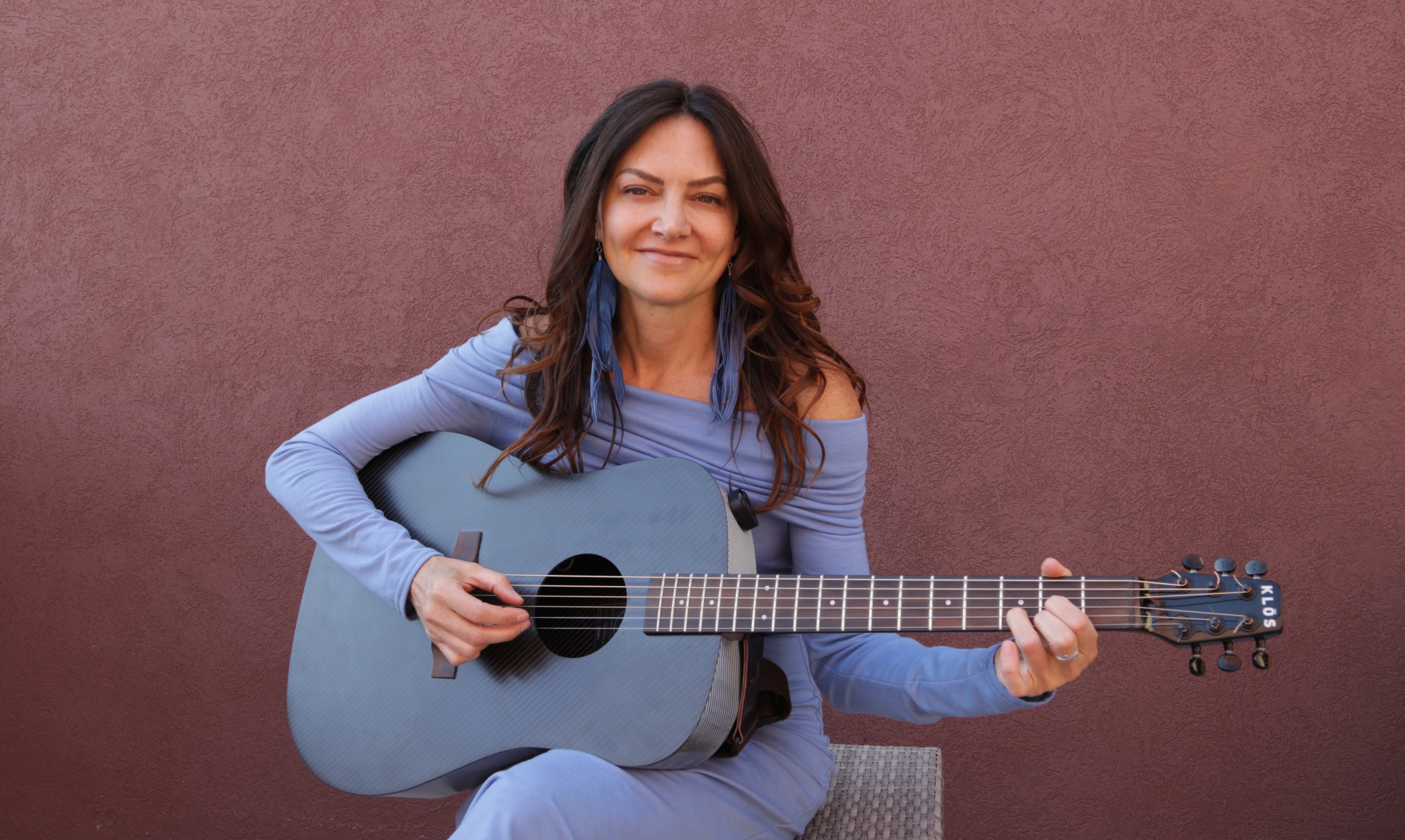 Woman with long dark hair holding a blue acoustic guitar, sitting against a plain background.