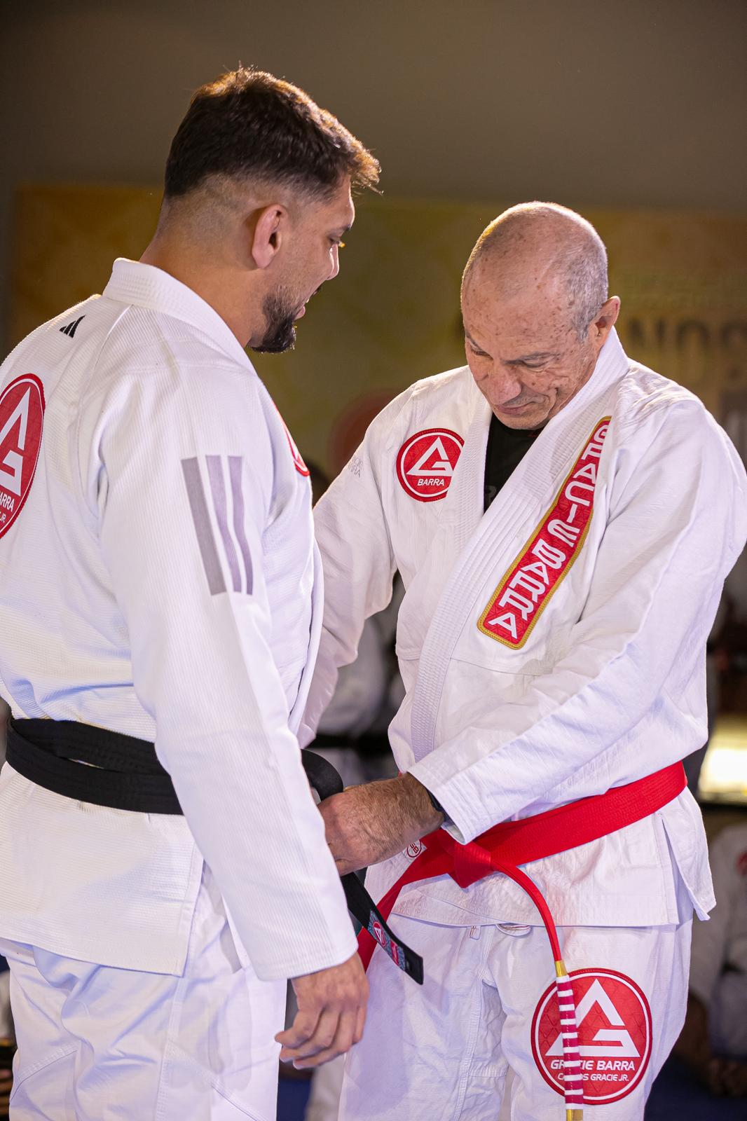 Two men in martial arts uniforms, one with a red belt, standing close and looking down, indoors with a plain background.