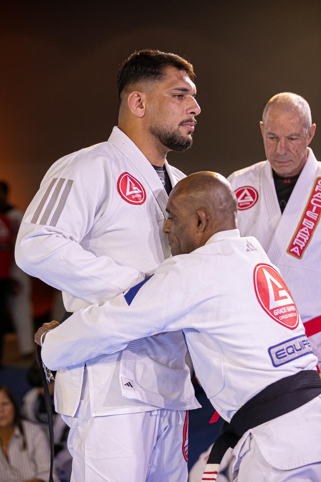 Two martial artists in white uniforms embrace, with a referee in the background, in a competition setting.