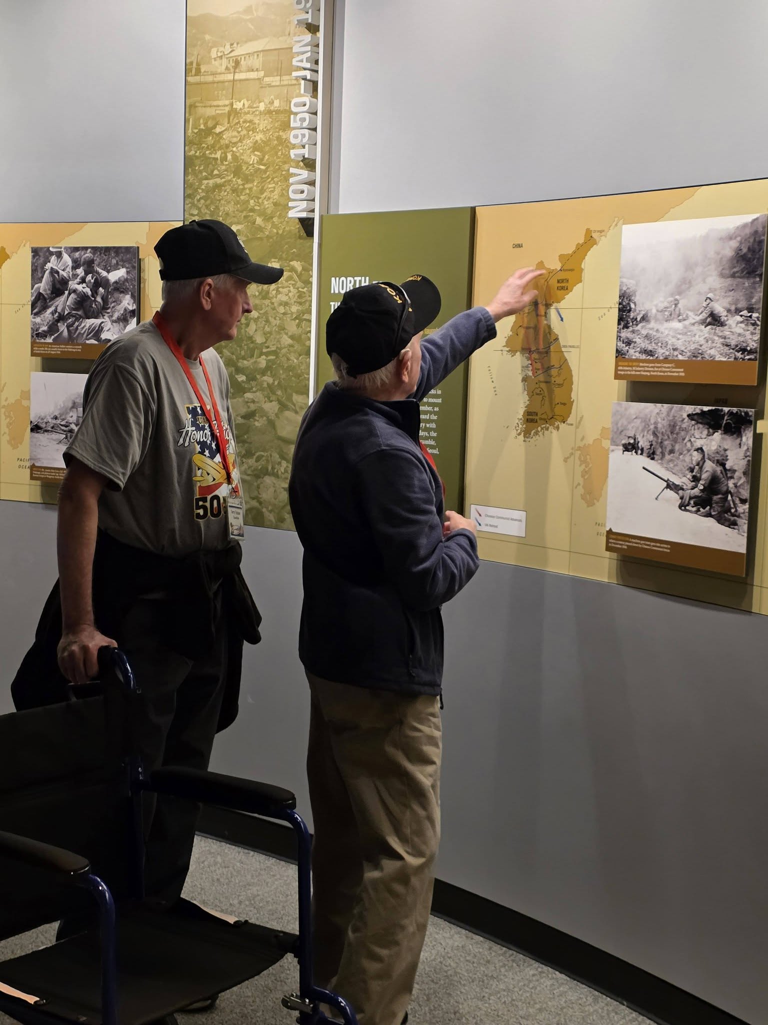 Two people look at photographs and a map on a display wall in an indoor exhibit.