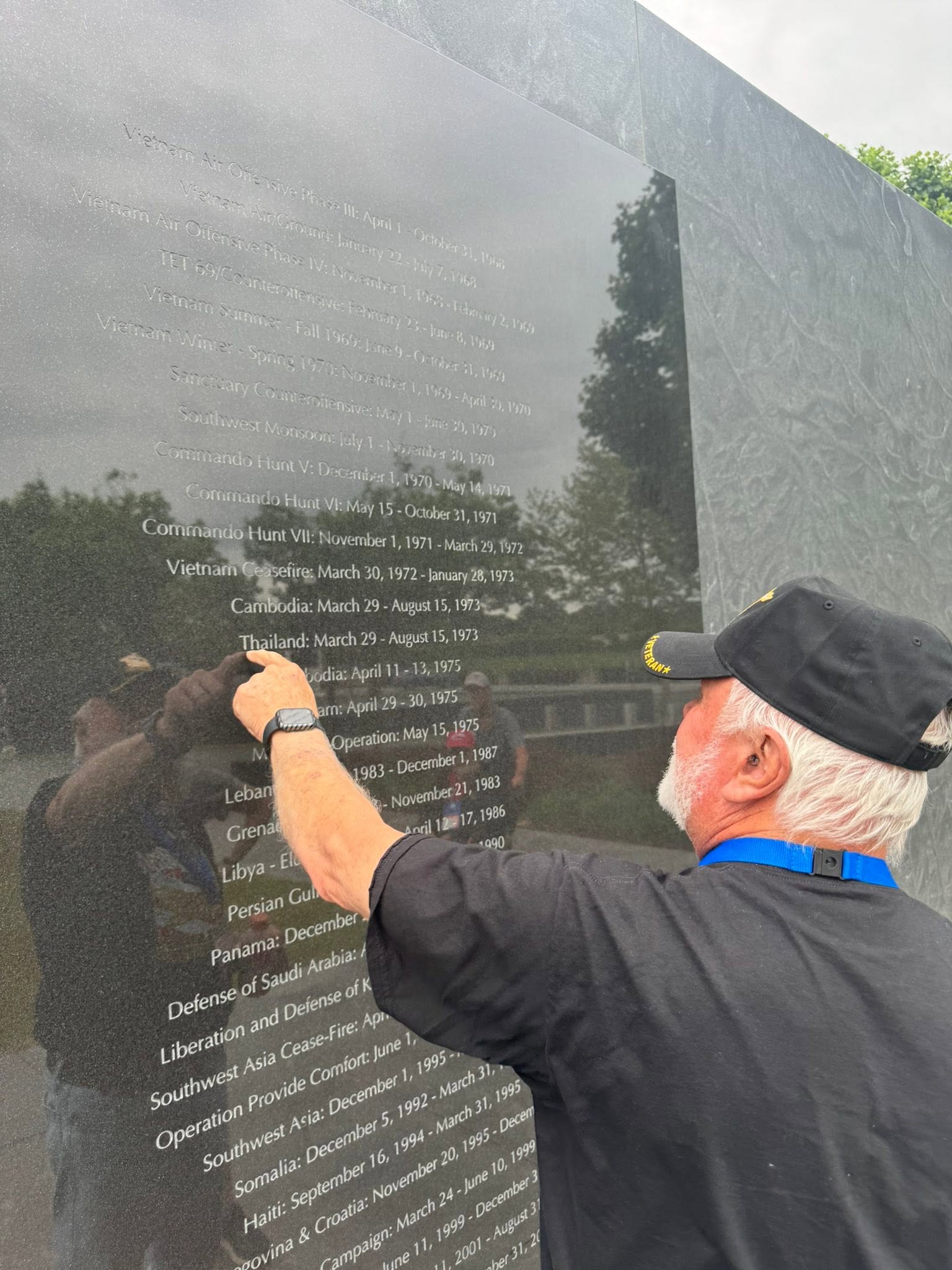 Older man with a black cap and glasses writing on a reflective black memorial wall, trees and sky reflected on the surface.