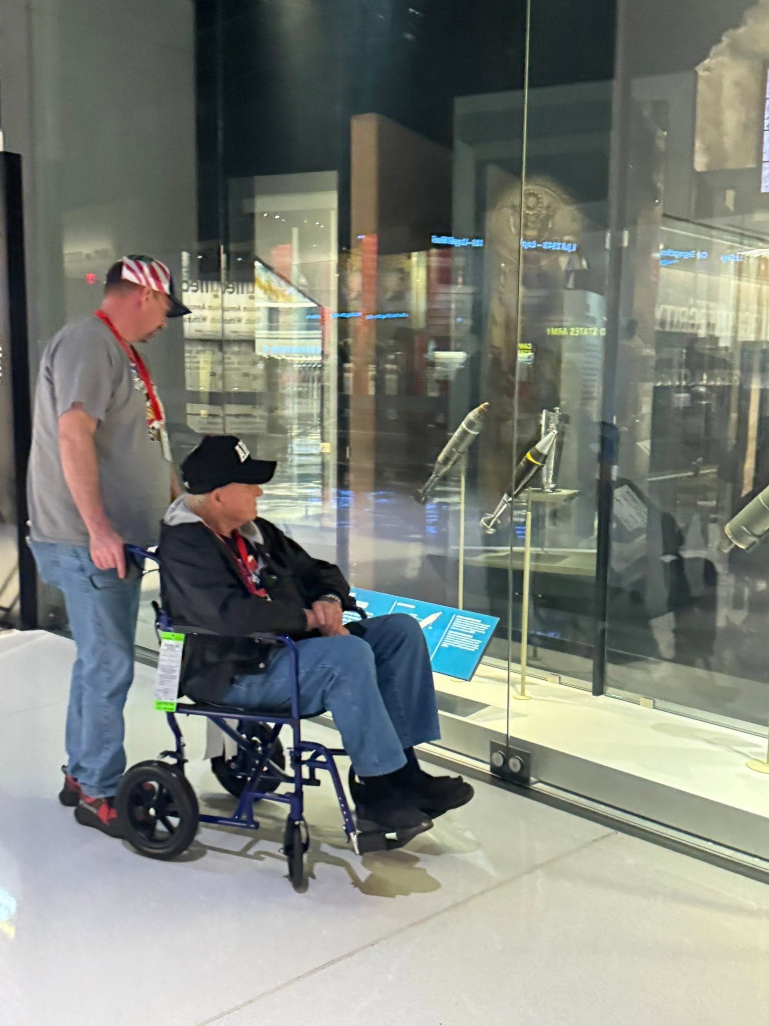 Two people, one in a wheelchair, looking at a display case with glass doors, inside a building with reflections on the glass.