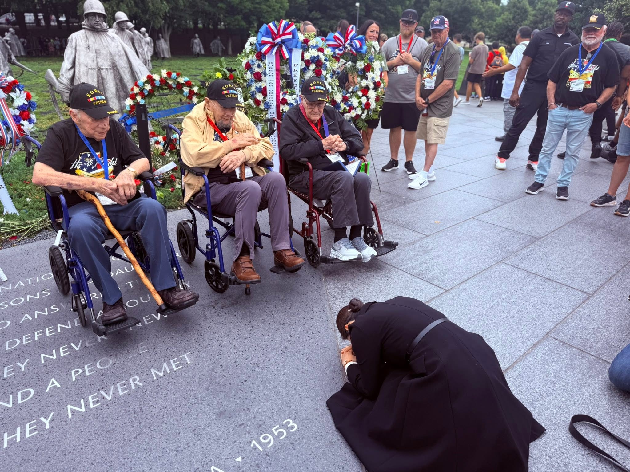 Three elderly people in wheelchairs at a memorial, with a person kneeling and others standing nearby, during a ceremony.
