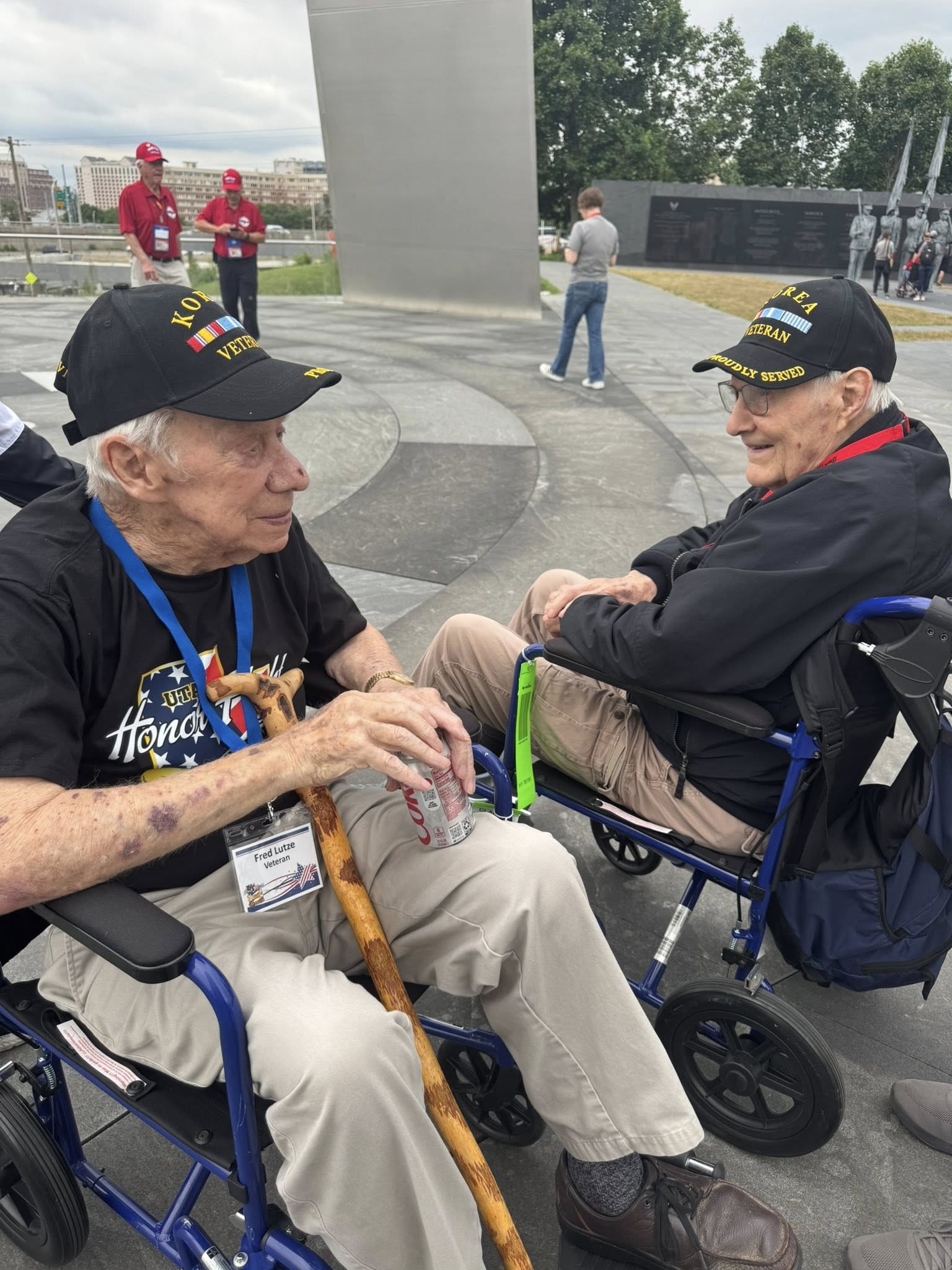 Two elderly men in wheelchairs wearing black hats and smiling, engaging in conversation outdoors with people in the background.