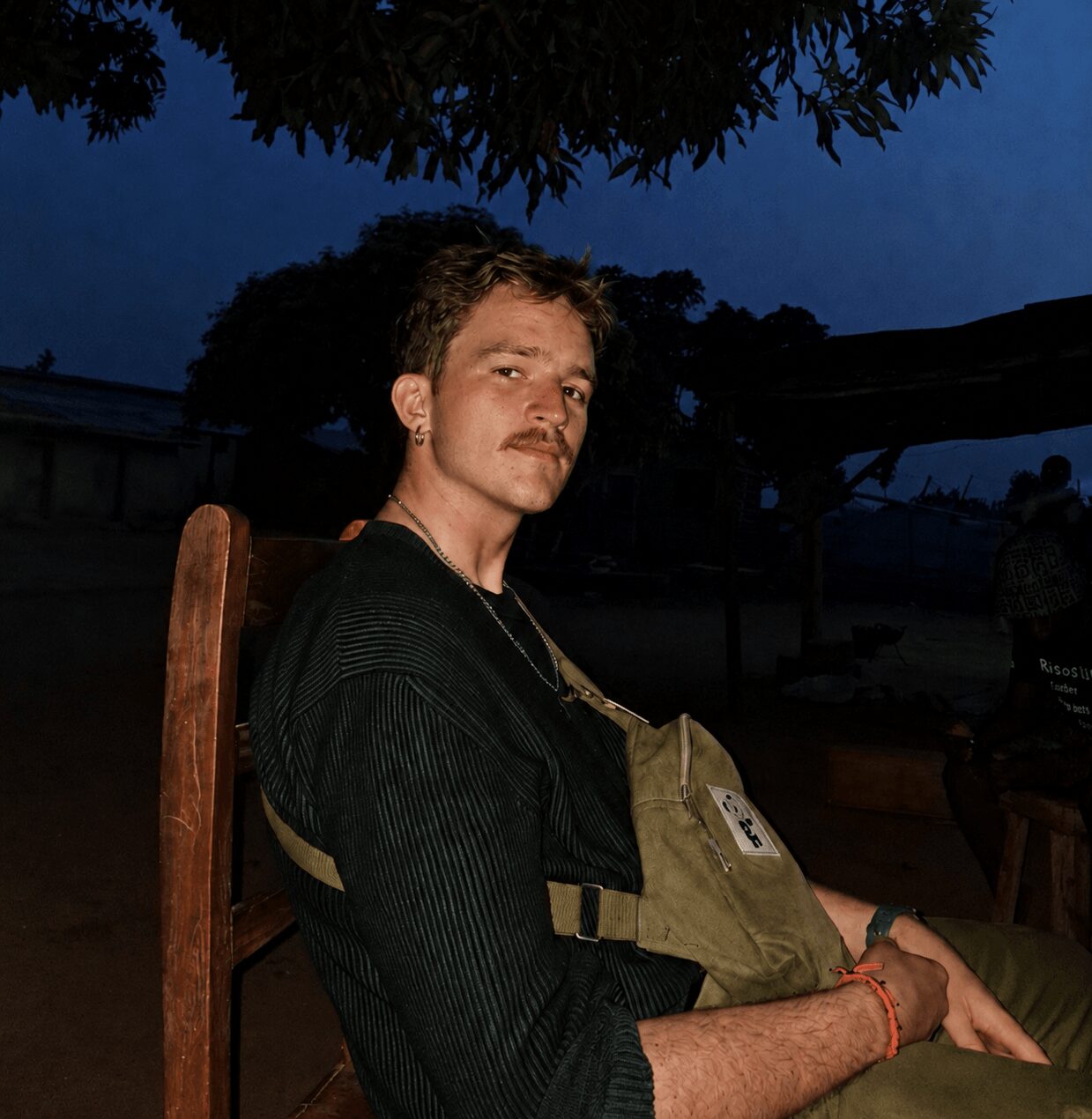Young man with light skin, short hair, and a mustache sitting outdoors at dusk, wearing a dark shirt and a bag.