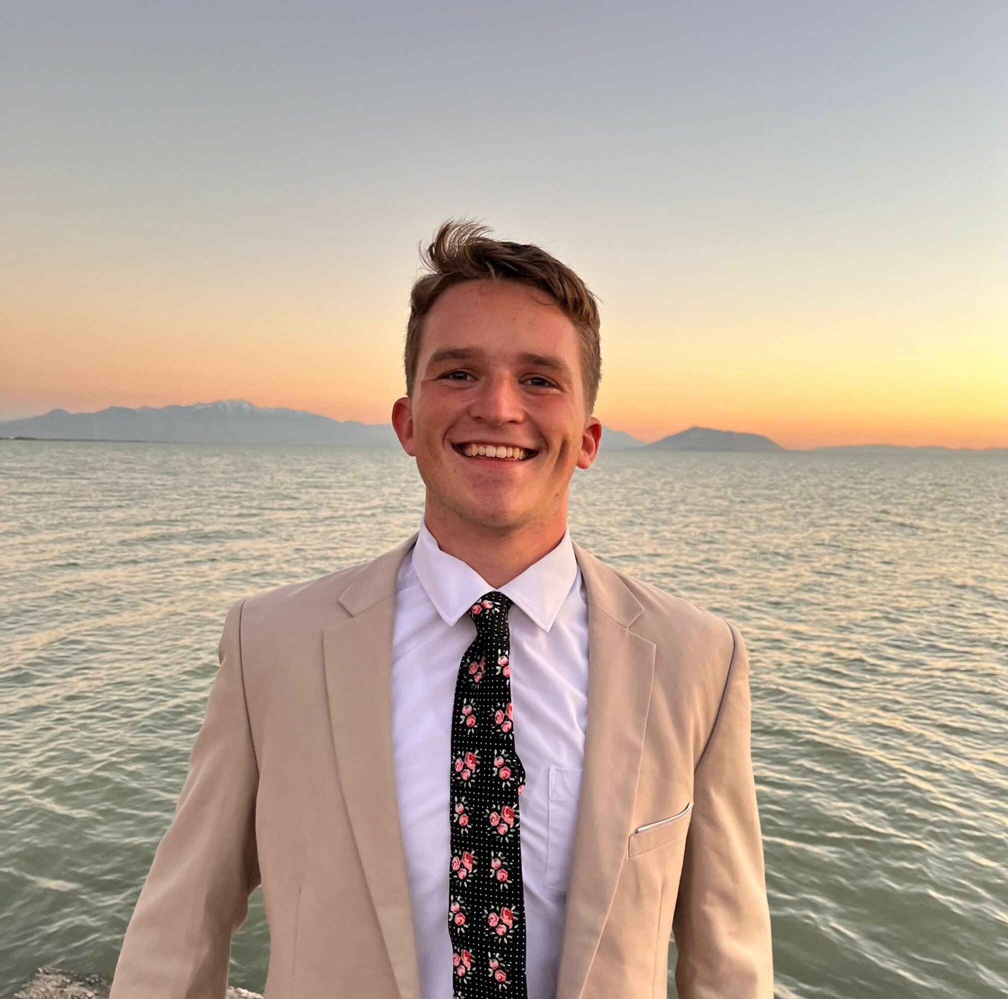 Young man smiling outdoors at sunset, wearing a beige blazer and a patterned tie, with water and mountains in background.