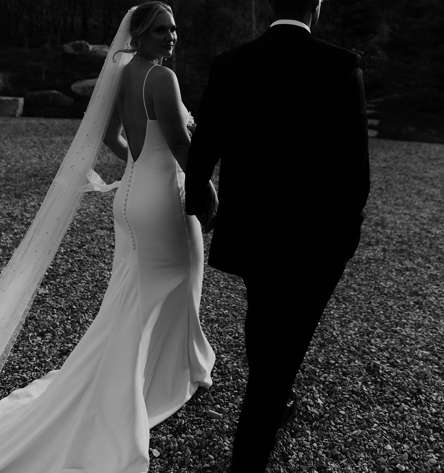 A bride and groom walk outdoors, holding hands, with the bride in a long dress and veil, and the groom in a suit.