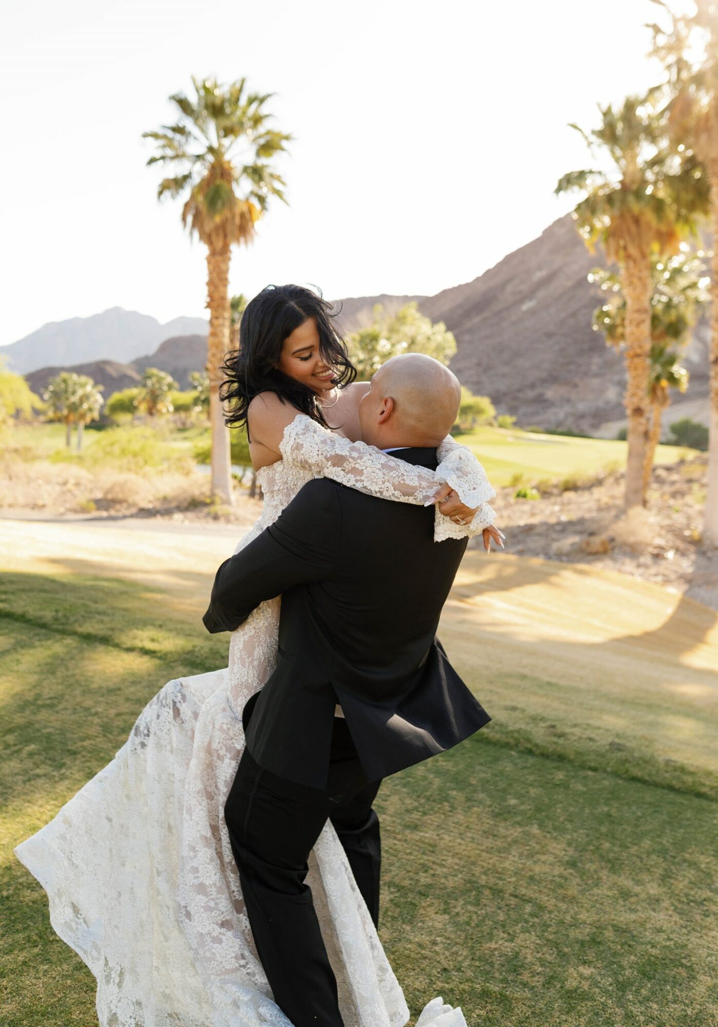 A couple embracing outdoors with palm trees and mountains in the background, woman in a white dress, man in a black suit.
