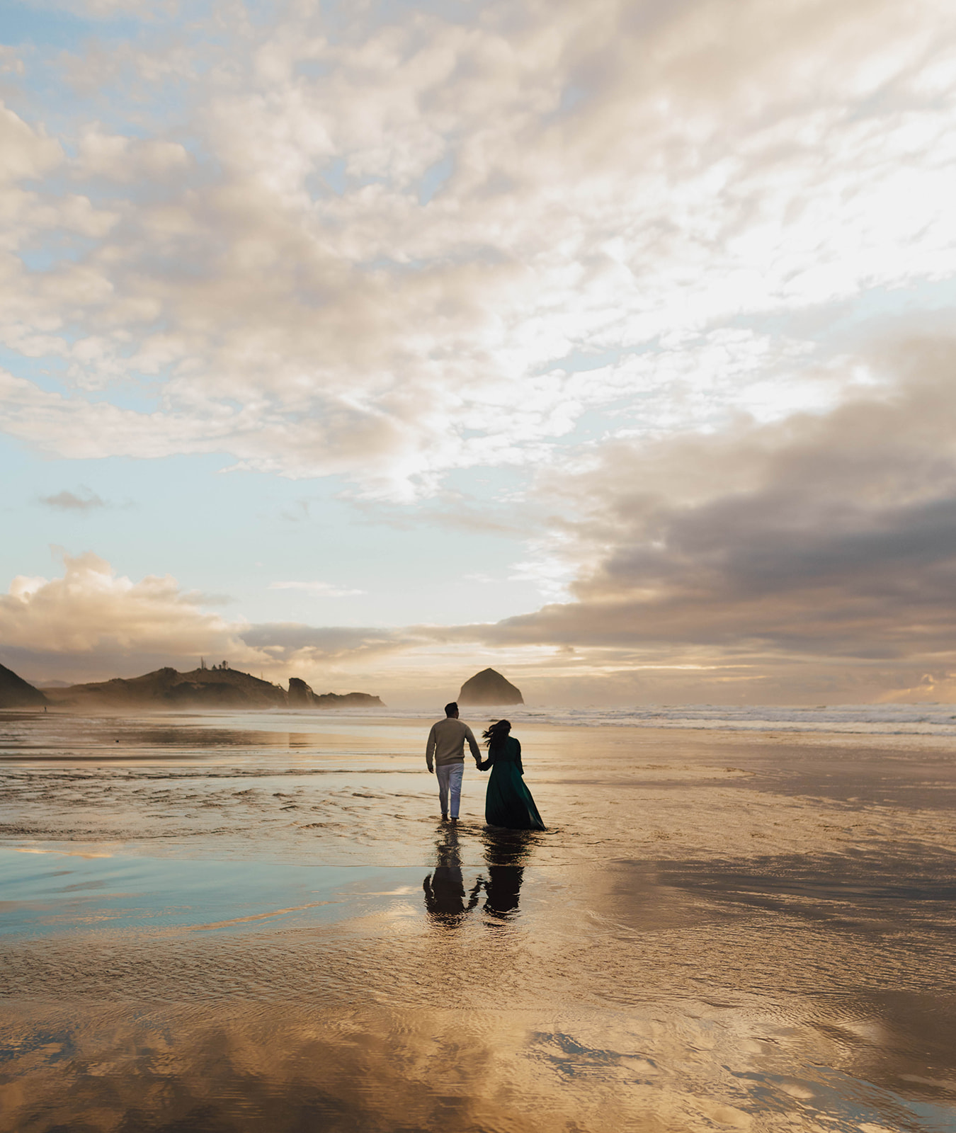 Two people stand on a beach with reflections in shallow water, clouds and rocks in the background, during sunset or sunrise.