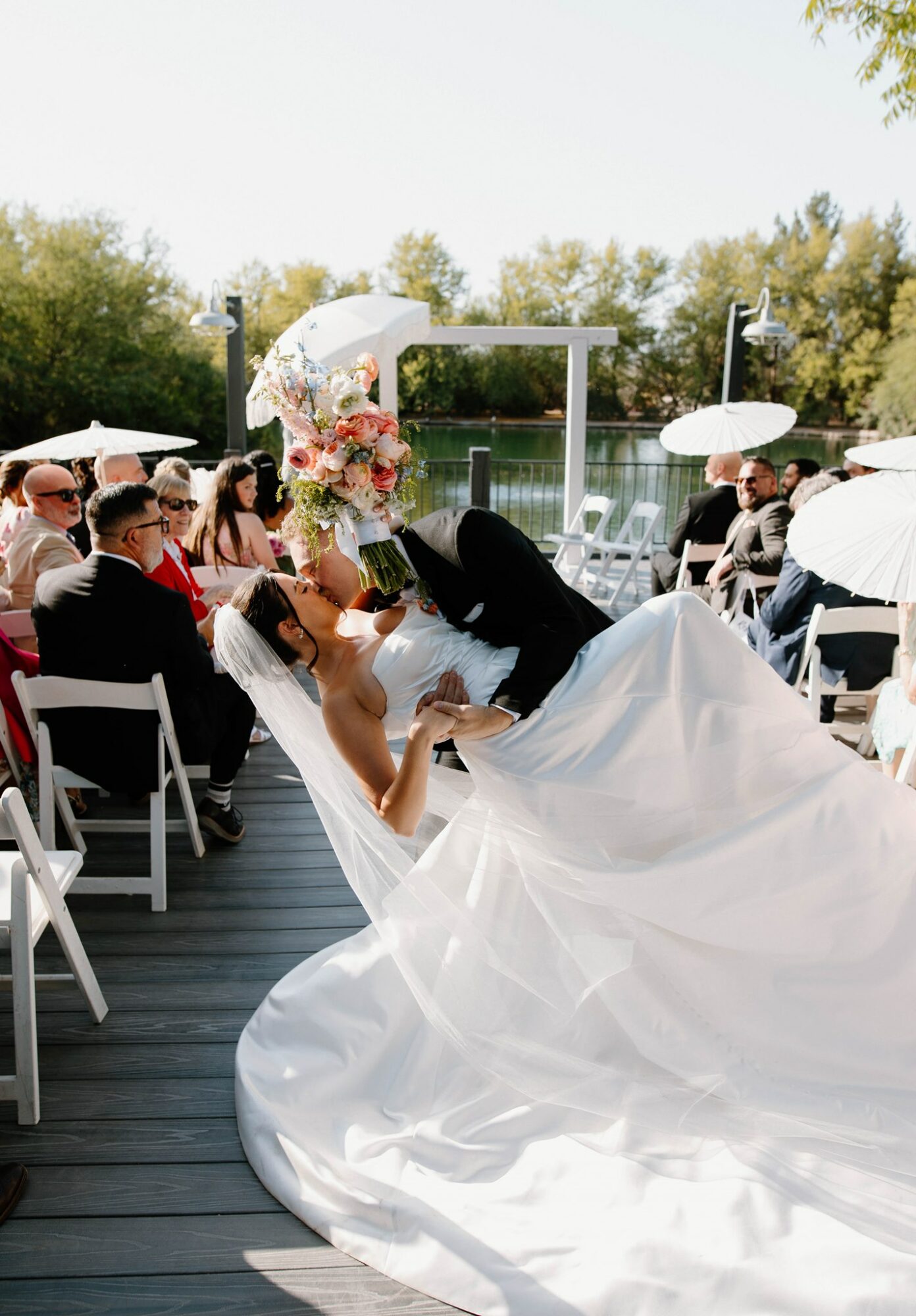 Bride and groom sharing a kiss outdoors during wedding ceremony, guests seated with umbrellas, trees in background.