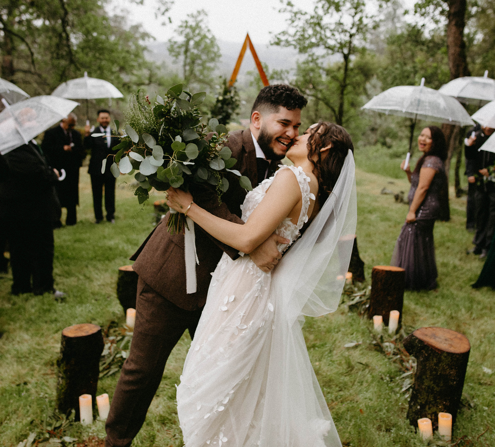 Couple dancing outdoors at wedding, surrounded by guests holding umbrellas, trees, and decorated with candles and logs.