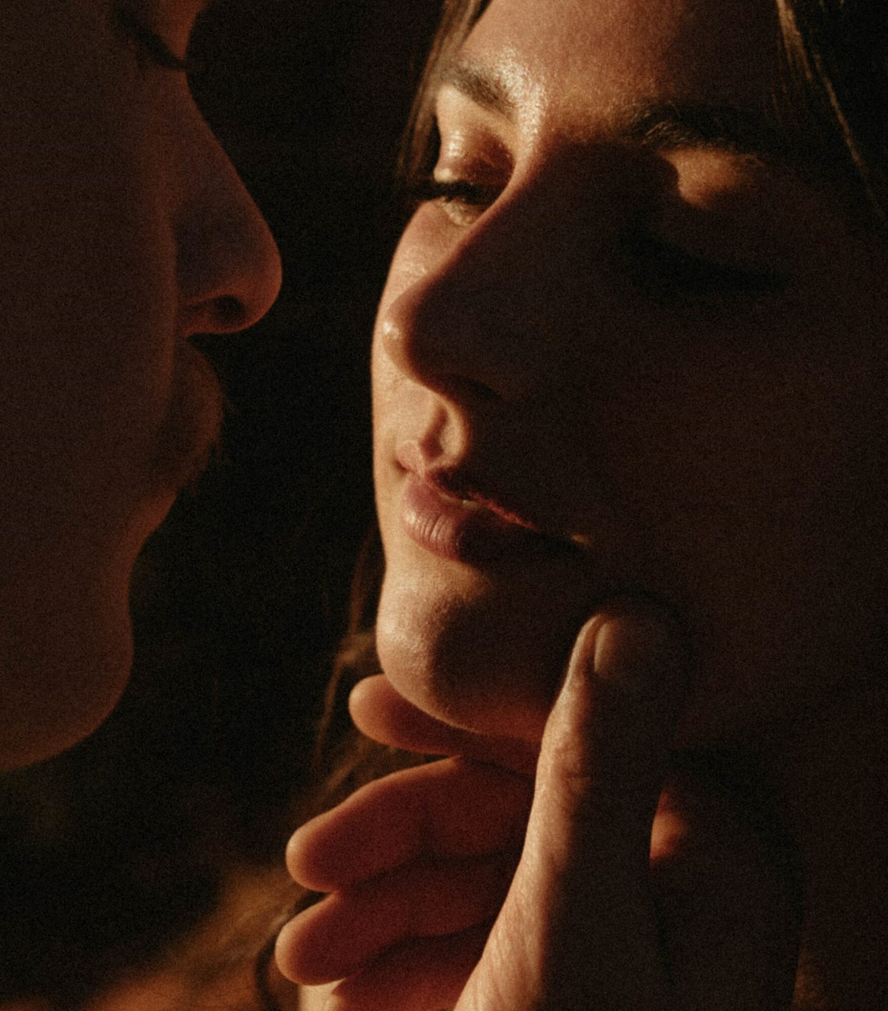 Close-up of a woman with eyes closed, touching her chin, with a person near her face in a dark setting.