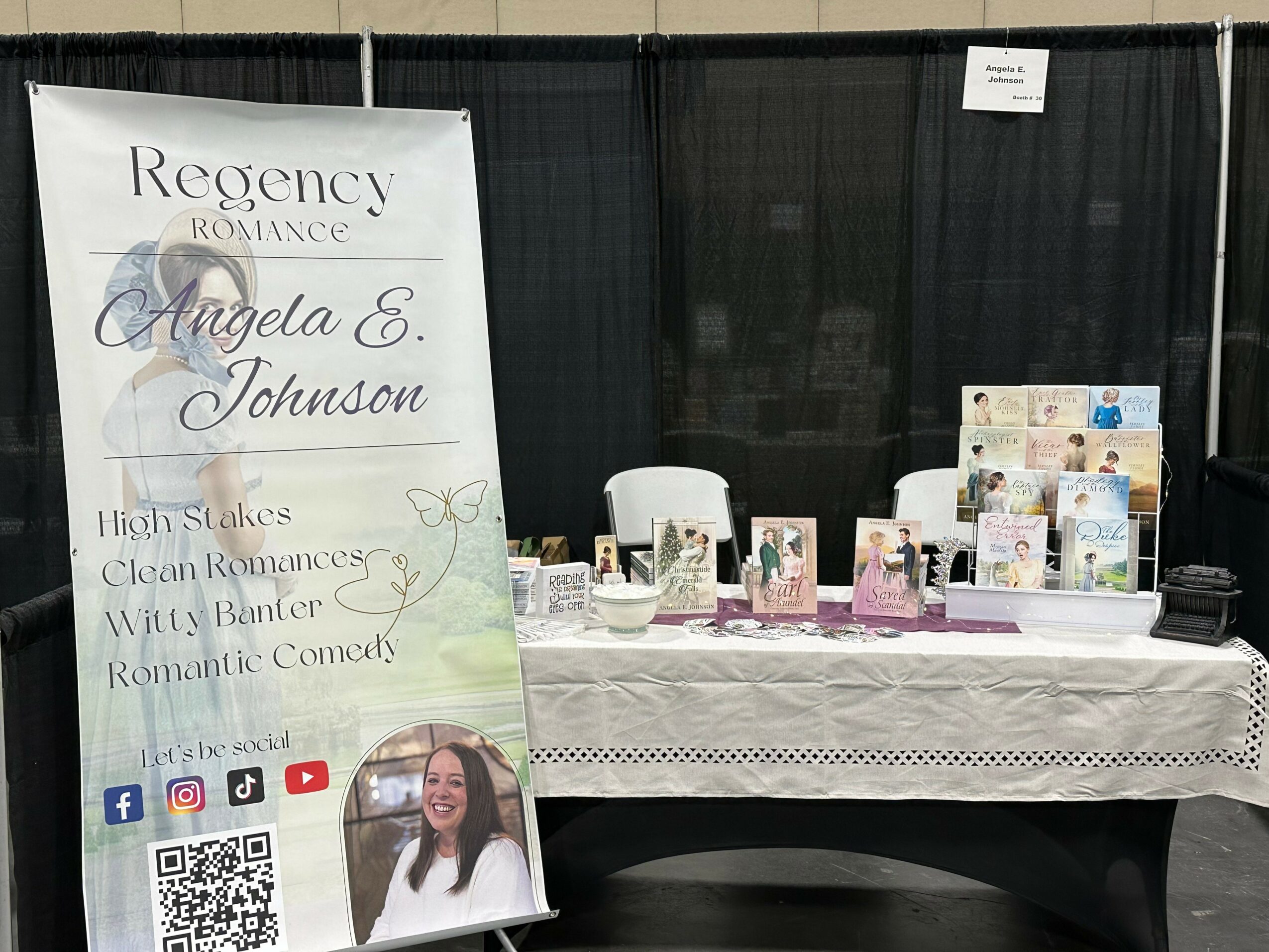 Book signing booth with a large sign for Angela E. Johnson's romance novels, table with books and promotional materials, black backdrop.