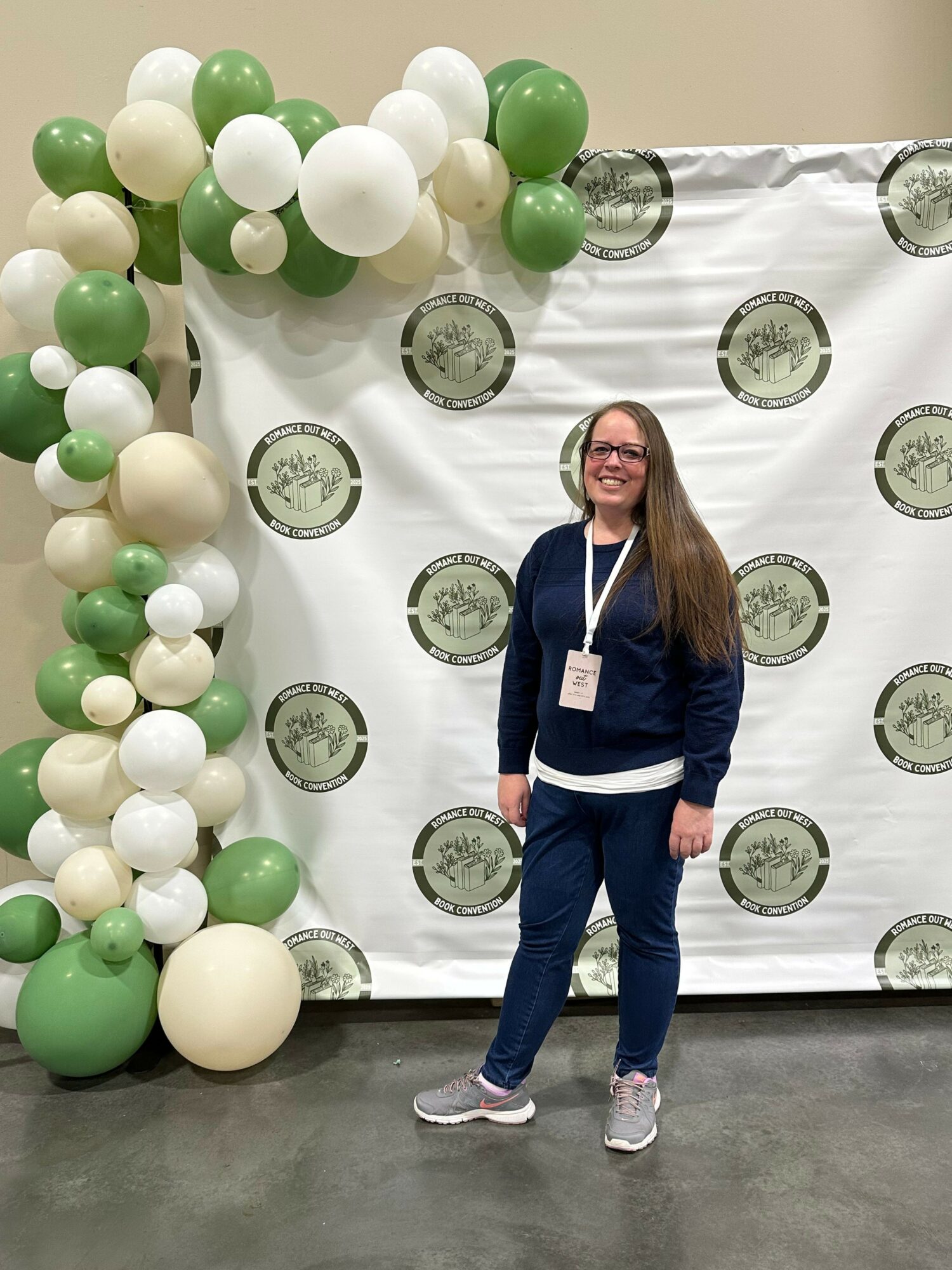 Woman standing next to balloon arrangement with white and green balloons, backdrop with circular logos, smiling, wearing a name badge.