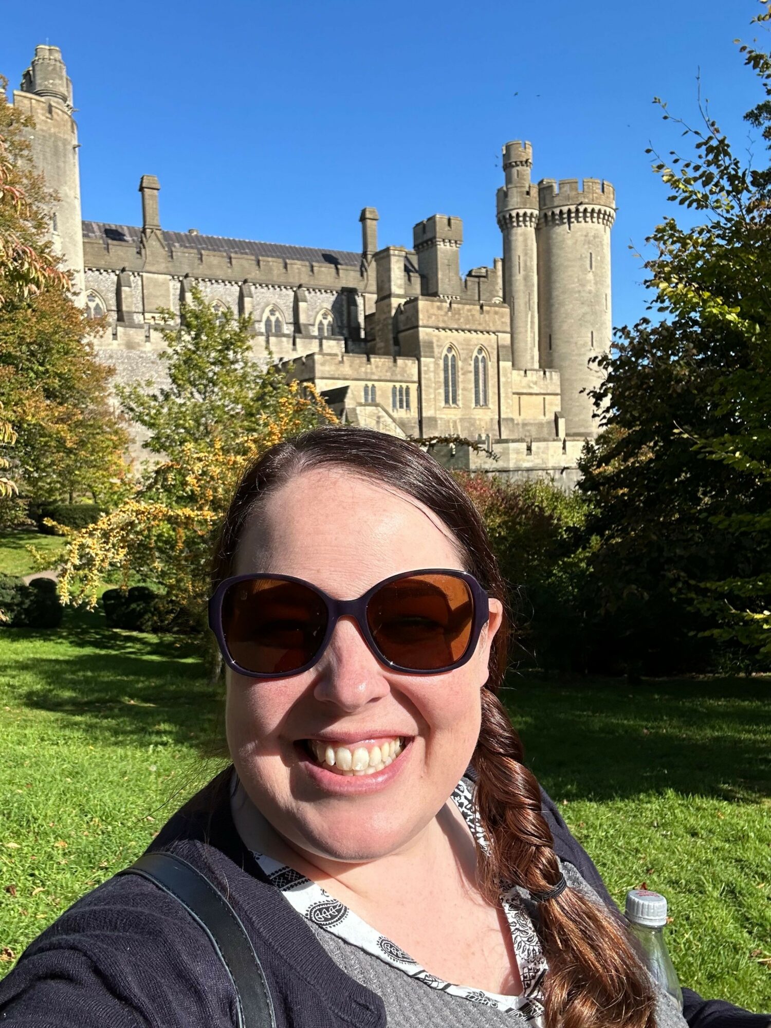 Young woman with sunglasses smiling outdoors near a castle with towers and a green lawn.