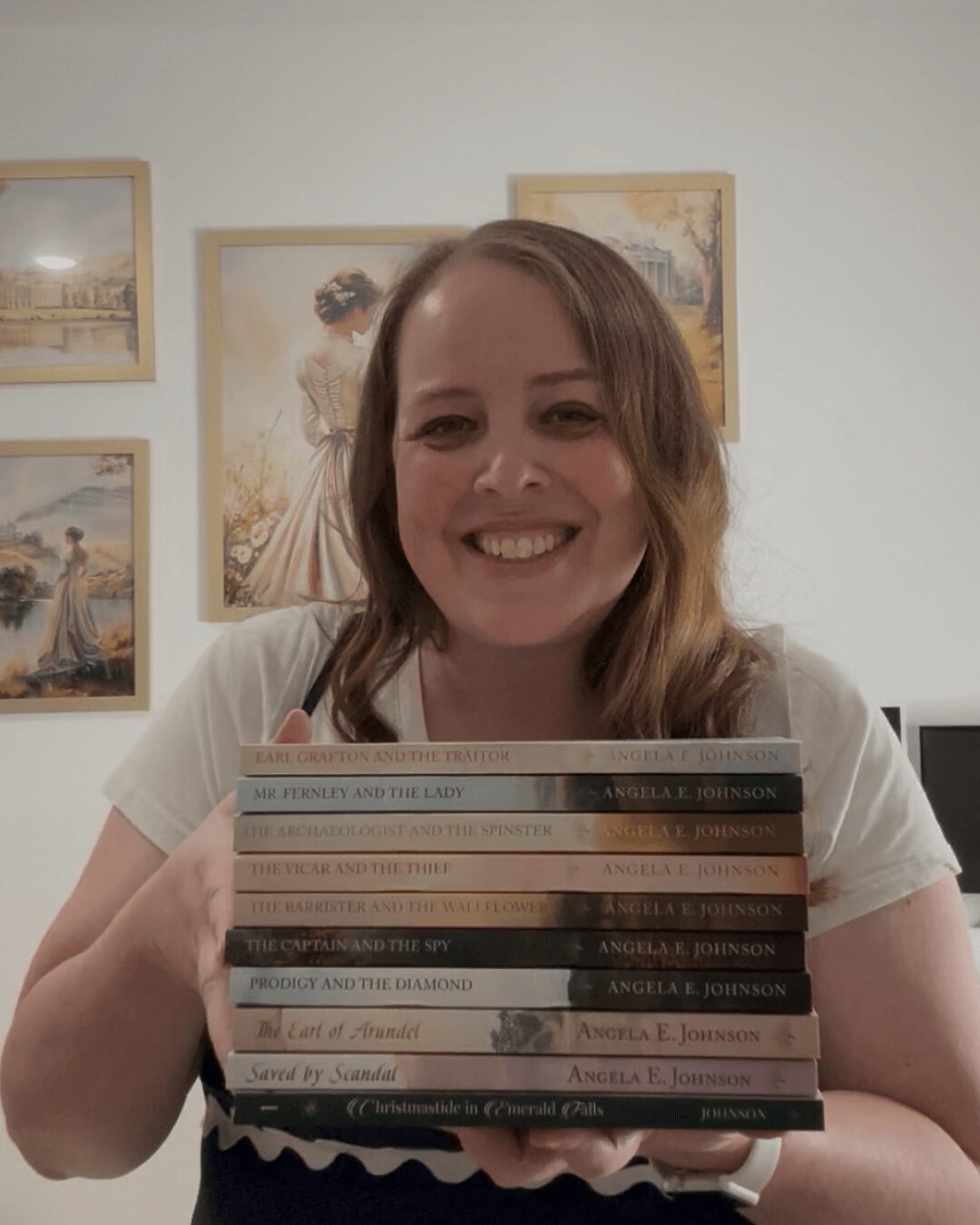 Young woman with shoulder-length hair smiling, holding a stack of books in front of a wall with framed pictures.
