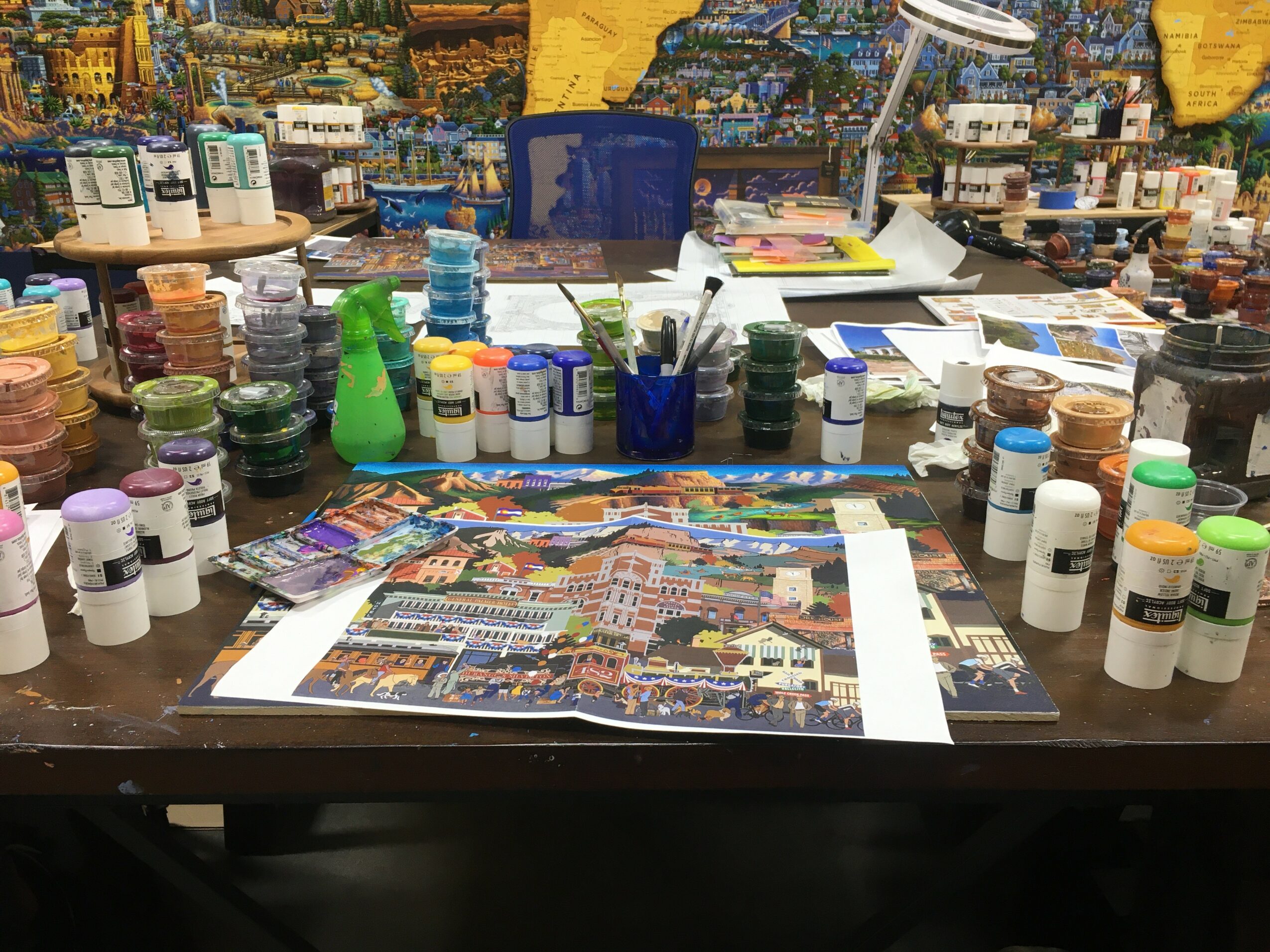 Art supplies, paints, brushes, and a colorful painting on a cluttered desk.