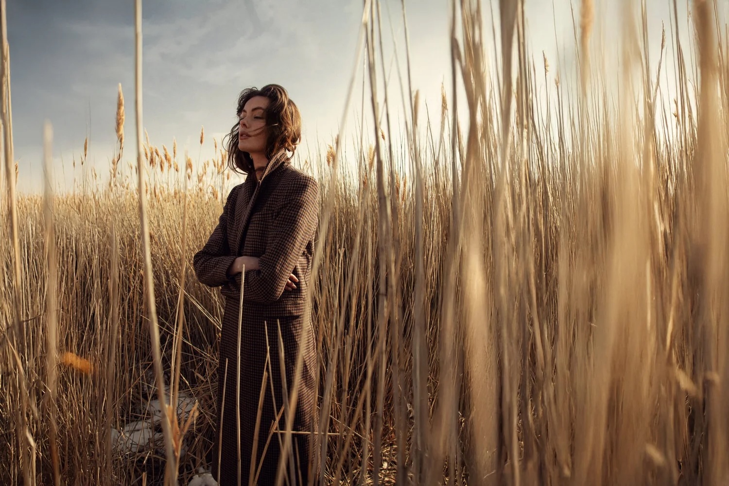 Woman standing in a field of tall grass during sunset, with arms crossed and smiling, wearing a dark jacket.