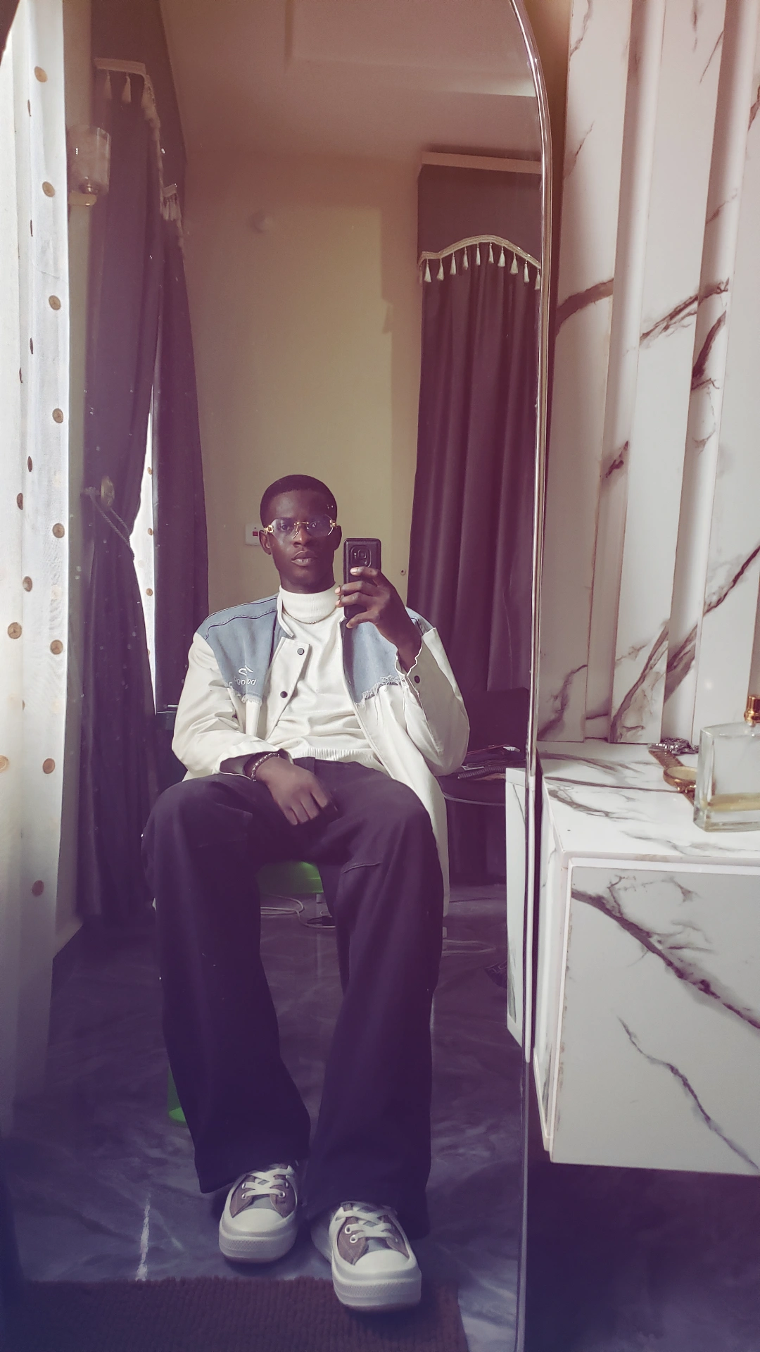 Person sitting on a chair taking a selfie in a room with curtains and a marble surface.