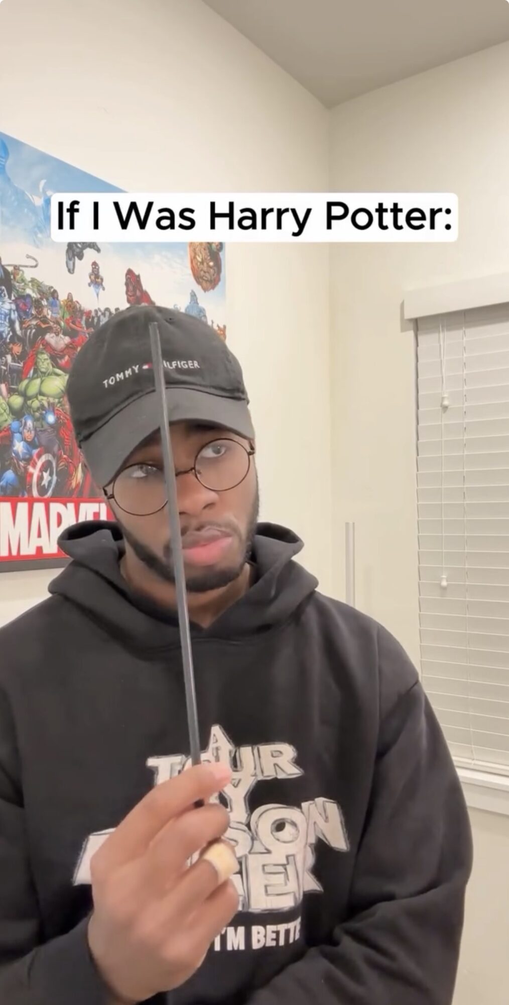 Person wearing glasses and a cap holding a wand in a room with a Marvel poster on the wall.