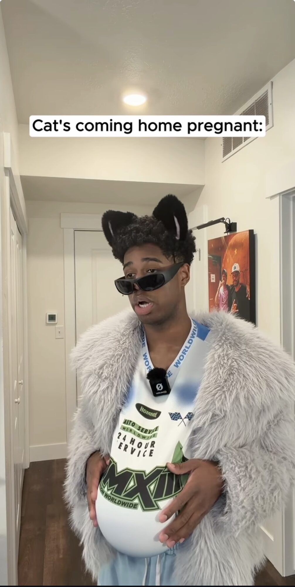 Person dressed as a cat with ears and sunglasses, holding a pregnant belly with a sign, in a hallway.