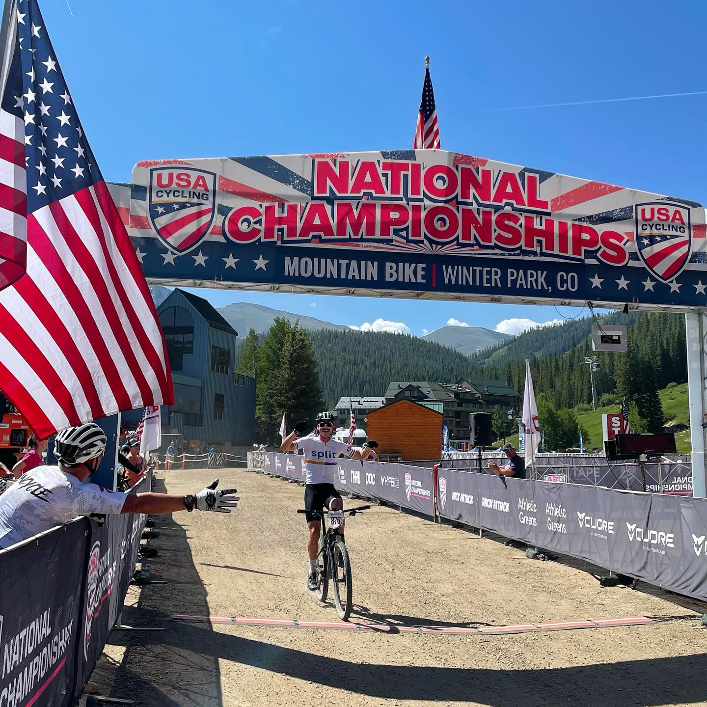 Cyclist crossing finish line at mountain bike race, with large banner overhead, flags, and spectators on side, in a mountain setting.