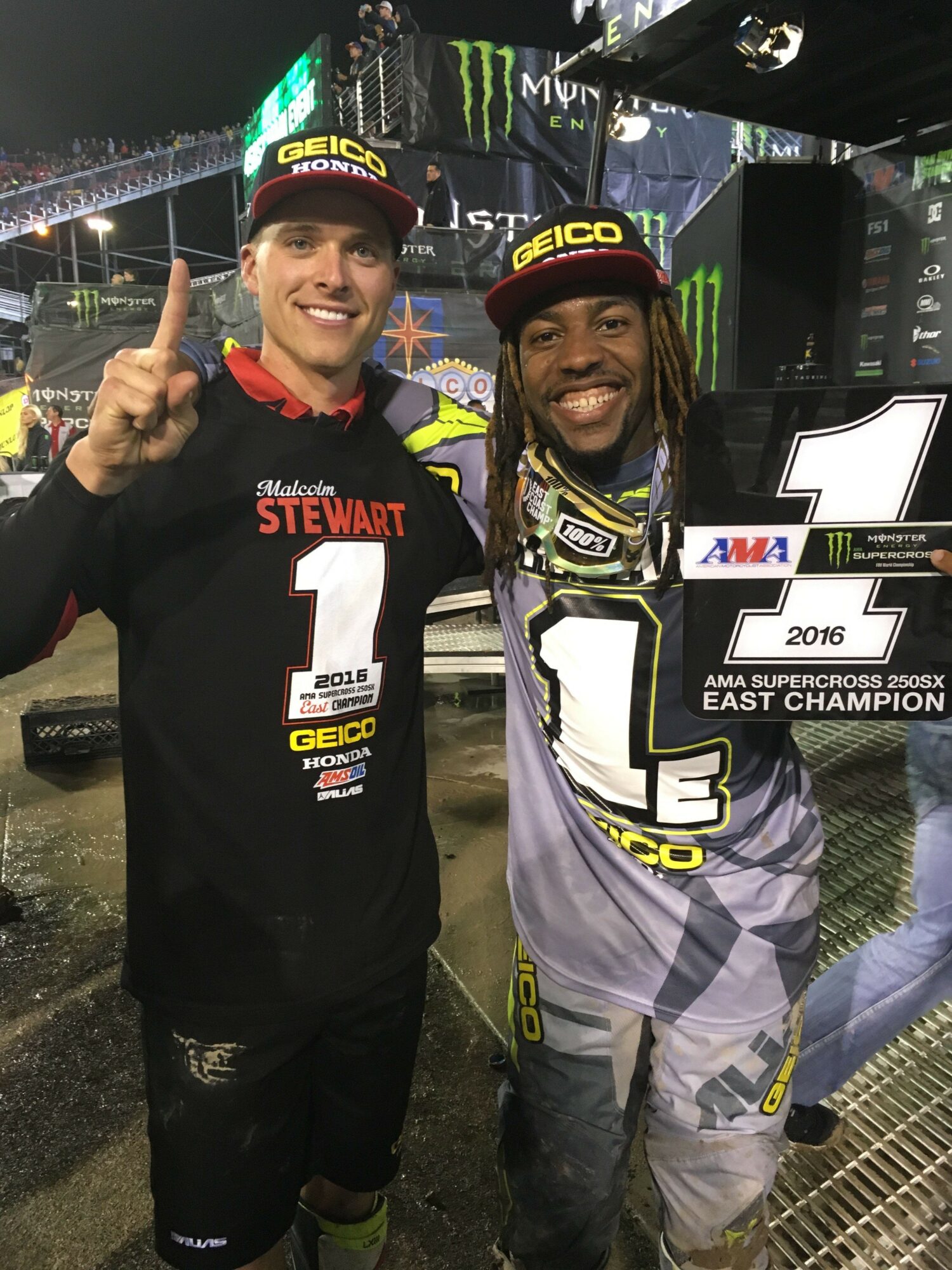 Two people celebrating, one holding a trophy, at a racing event with a race track background.