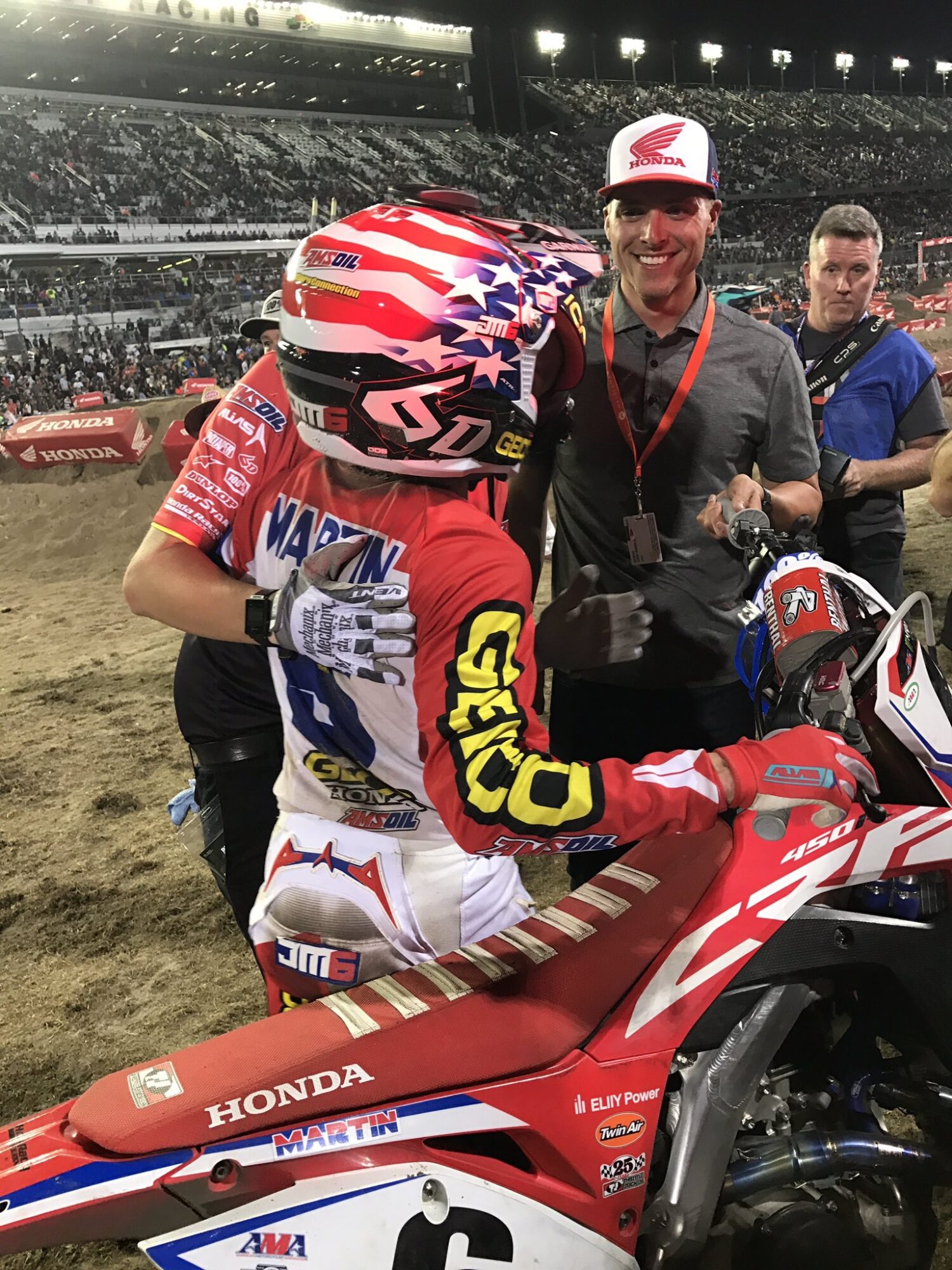Motocross rider in red, white, and blue gear with helmet, next to a red Honda dirt bike, in a stadium with spectators.
