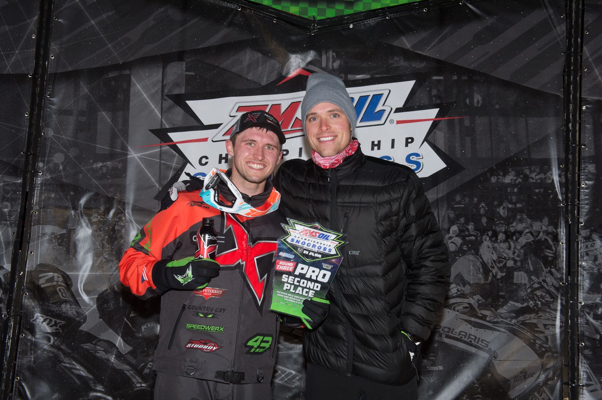 Two men smiling, one holding a trophy, standing in front of a racing championship backdrop, wearing jackets and hats.