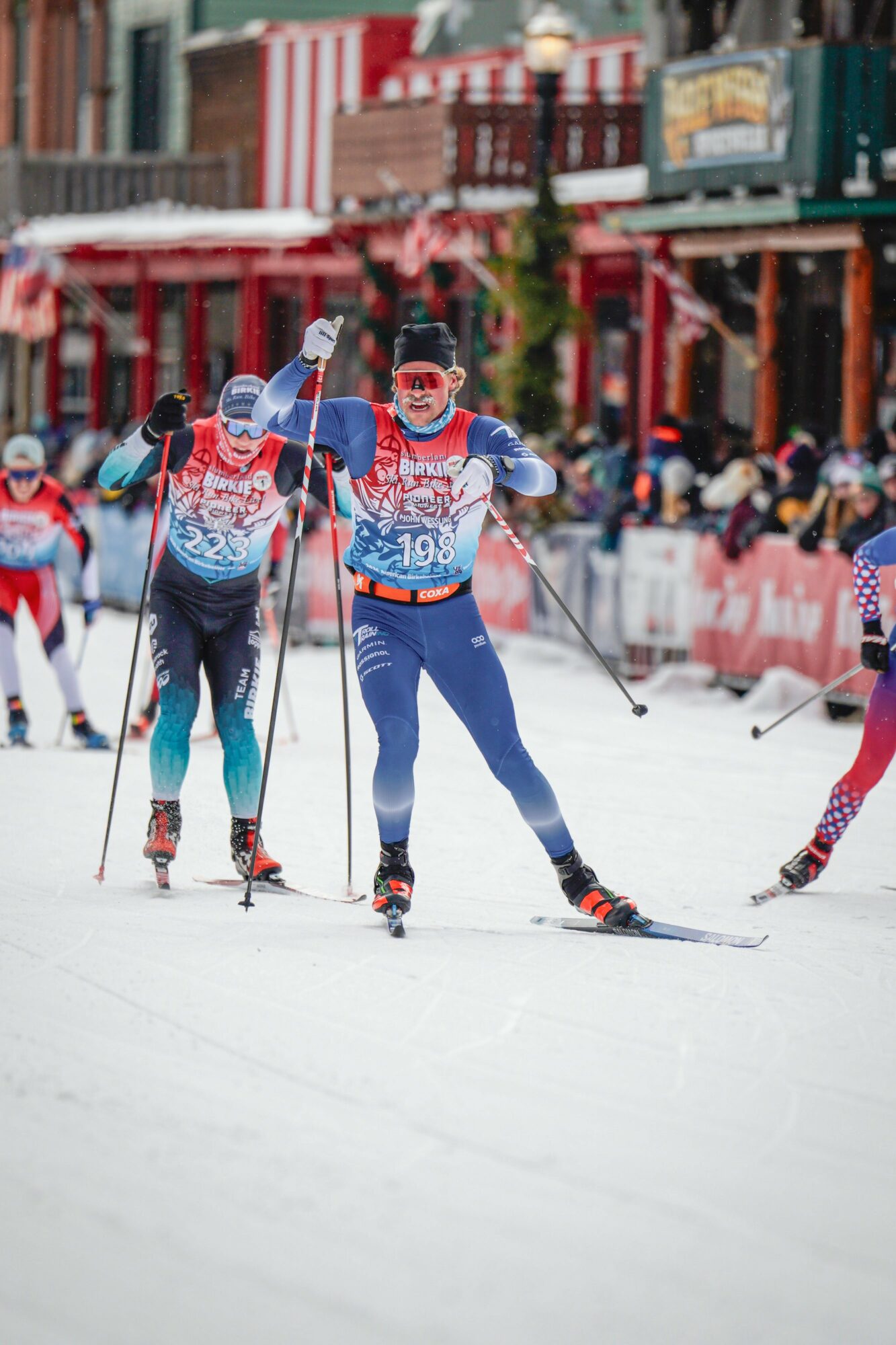 Group of cross-country skiers racing on snow with colorful buildings in background.