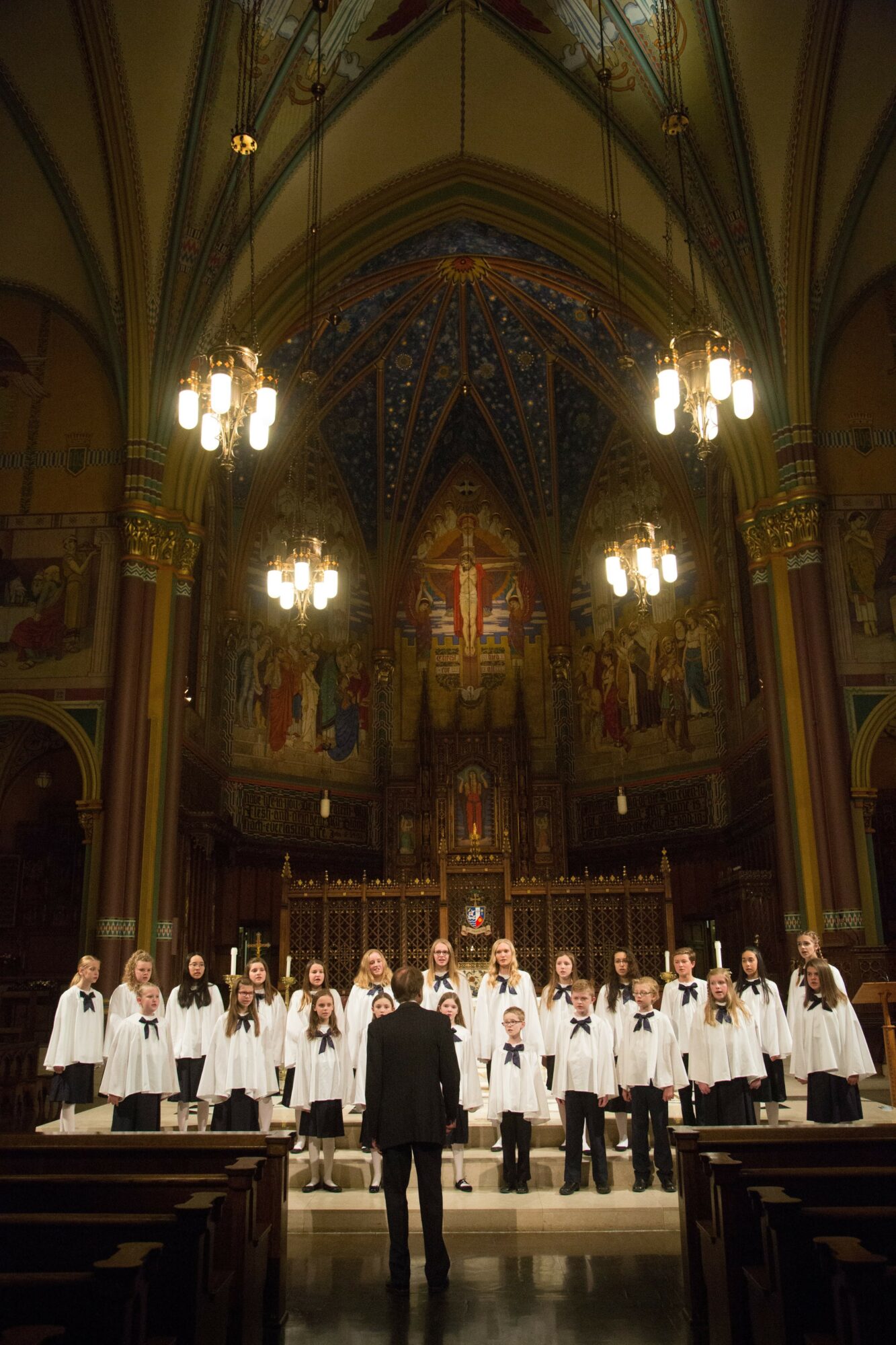 Choir of children in white robes performs in a church with ornate architecture and stained glass windows, conductor in front.