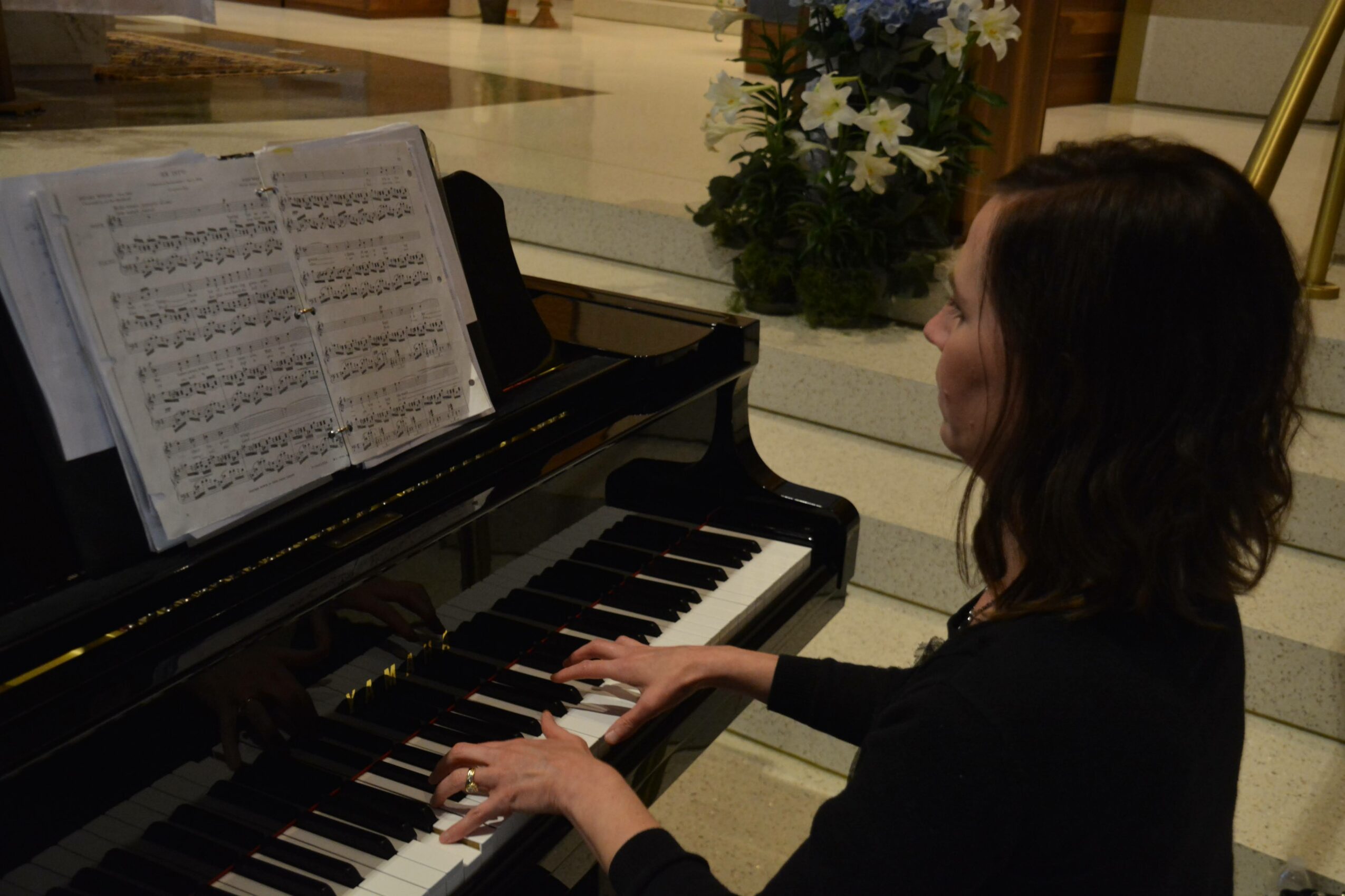 Woman playing piano with sheet music open, in a well-lit indoor space, with plants in the background.