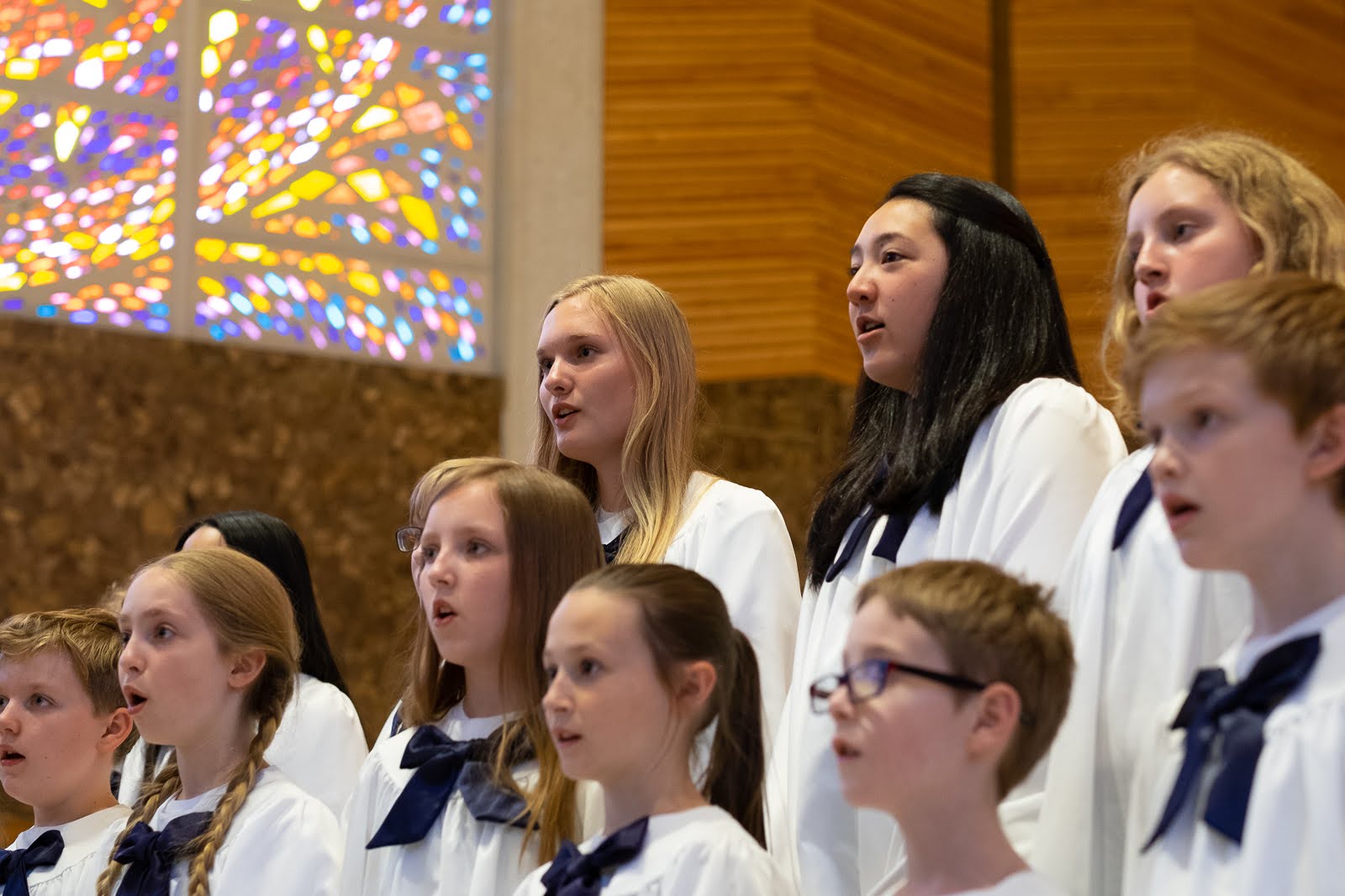 Group of children singing in choir, inside a church with stained glass window, wooden walls, and choir robes.