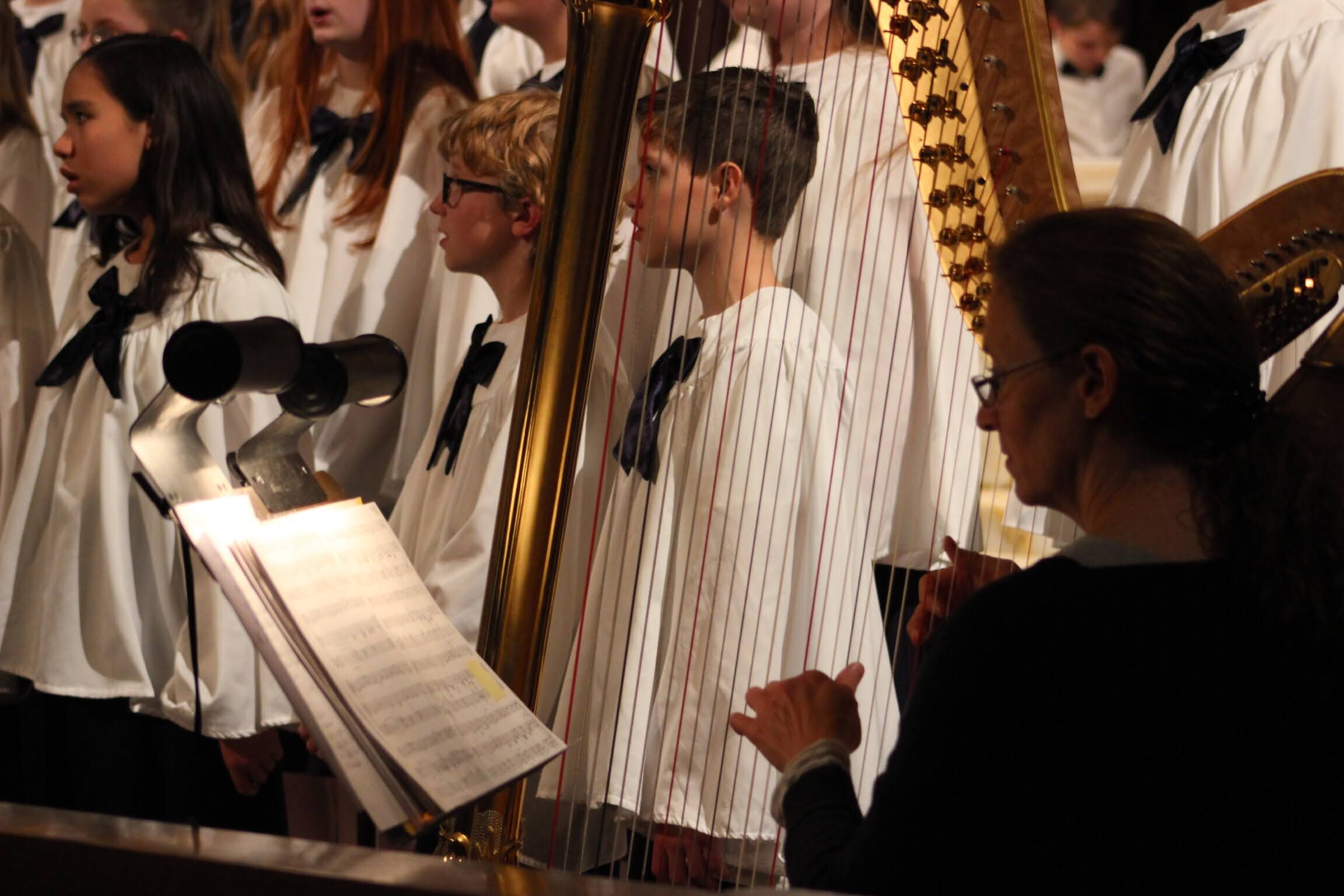 Group of children singing in choir with a conductor, music stand, and harp visible, in a performance setting.
