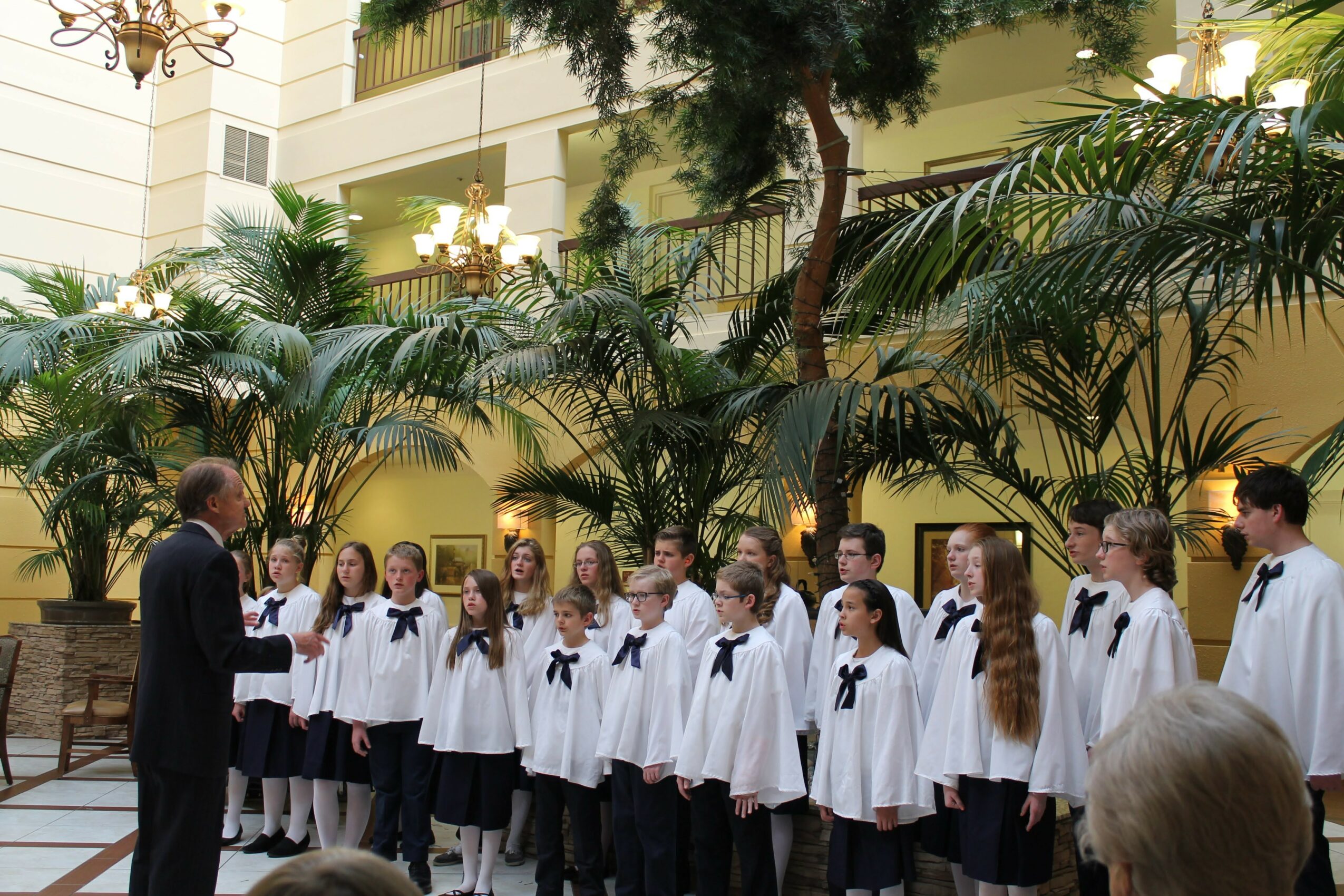 Group of children in white shirts and black bows standing in indoor garden, listening to a person speaking, with lush plants and yellow walls.