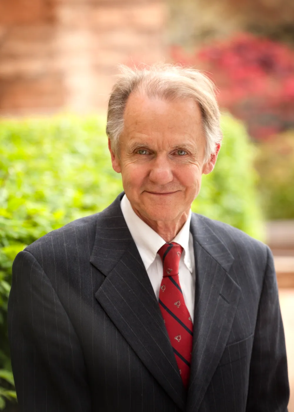 Older man in a suit and tie outdoors with greenery and blurred background, smiling.