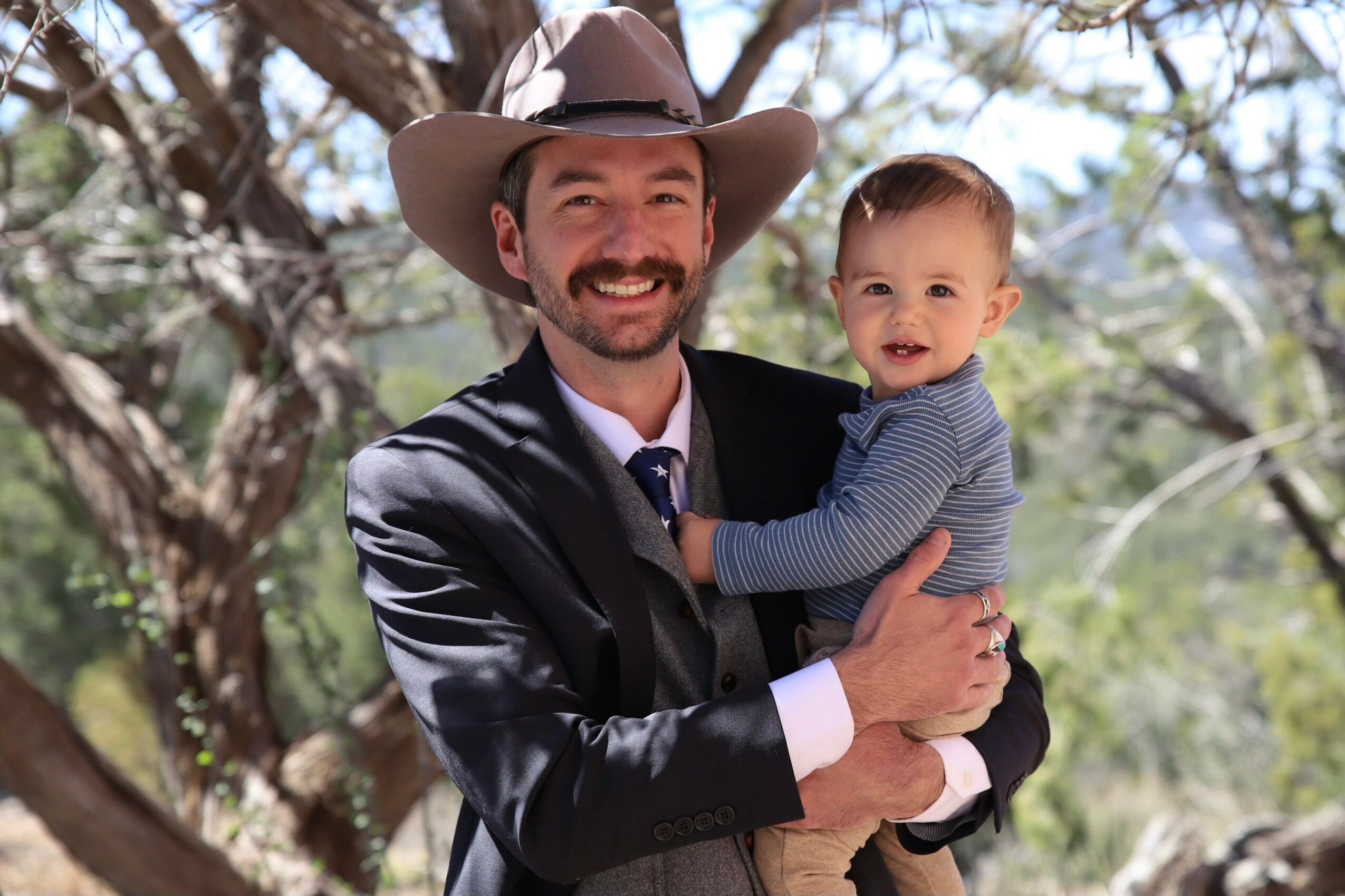 Man in a suit and cowboy hat holding a smiling baby outdoors with trees in the background.