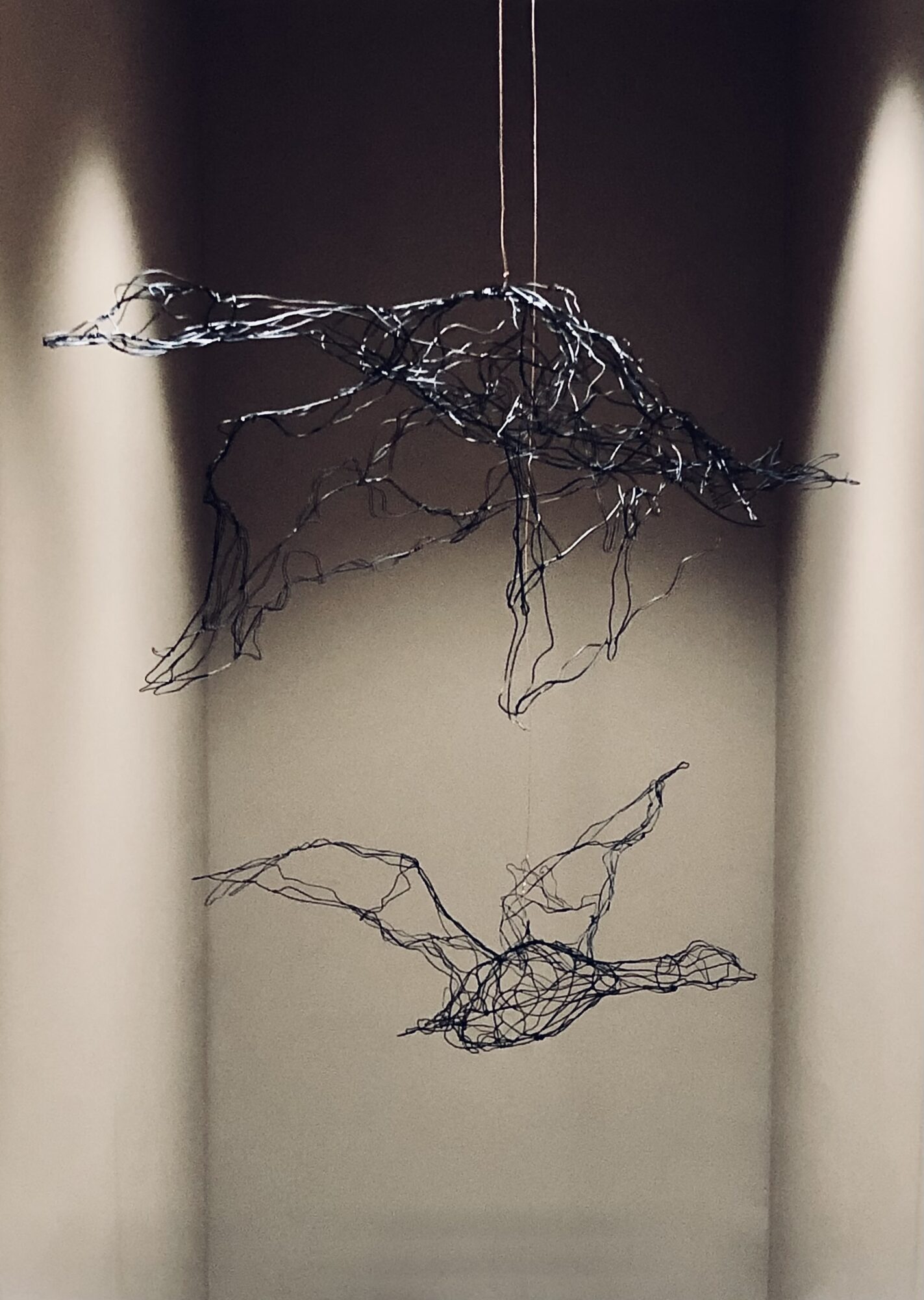 Wire sculpture hanging from ceiling, with abstract, tangled shapes, against a dark background with light walls.