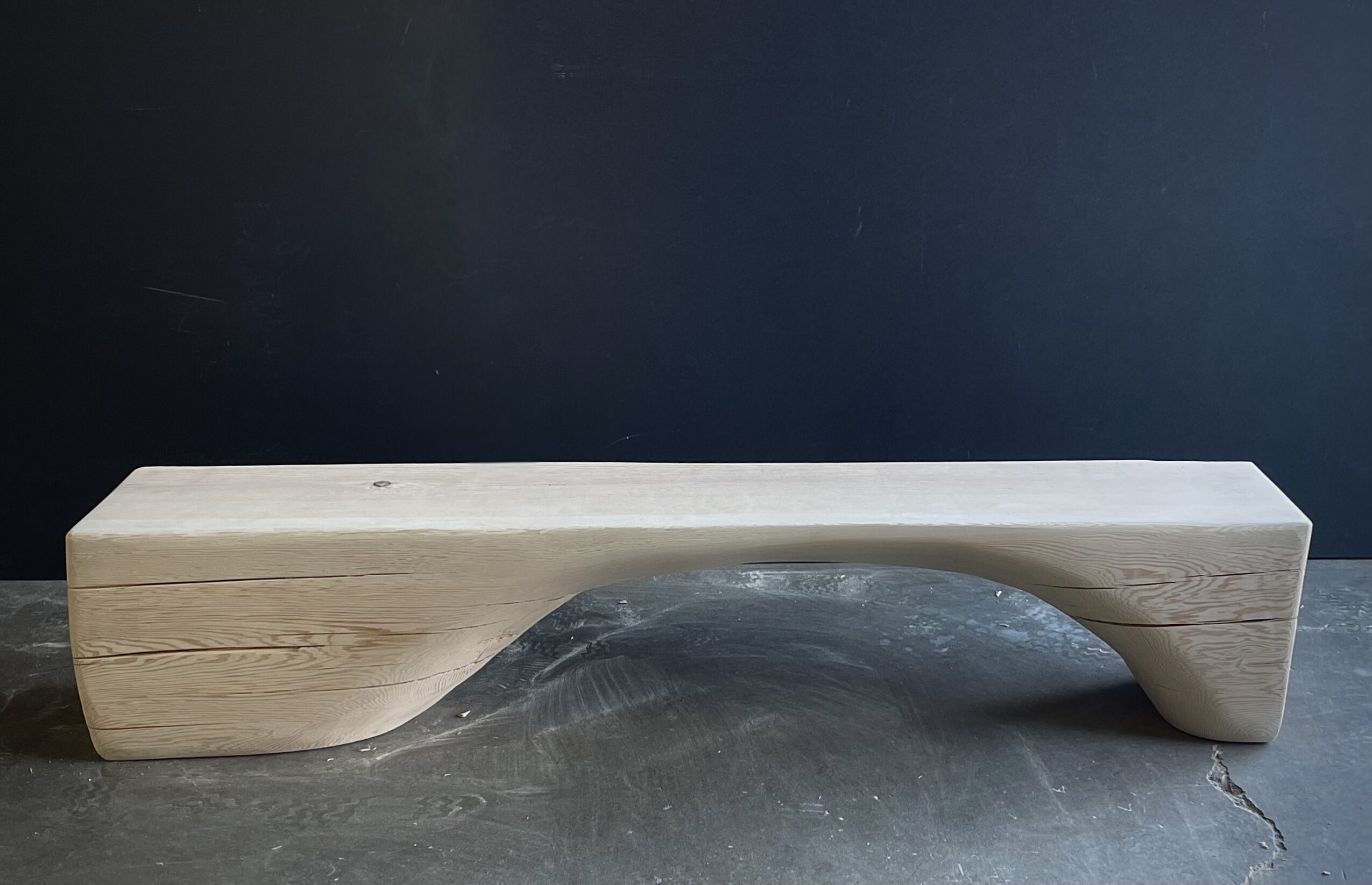 Long wooden bench with a curved cutout in the middle, placed against a dark wall.