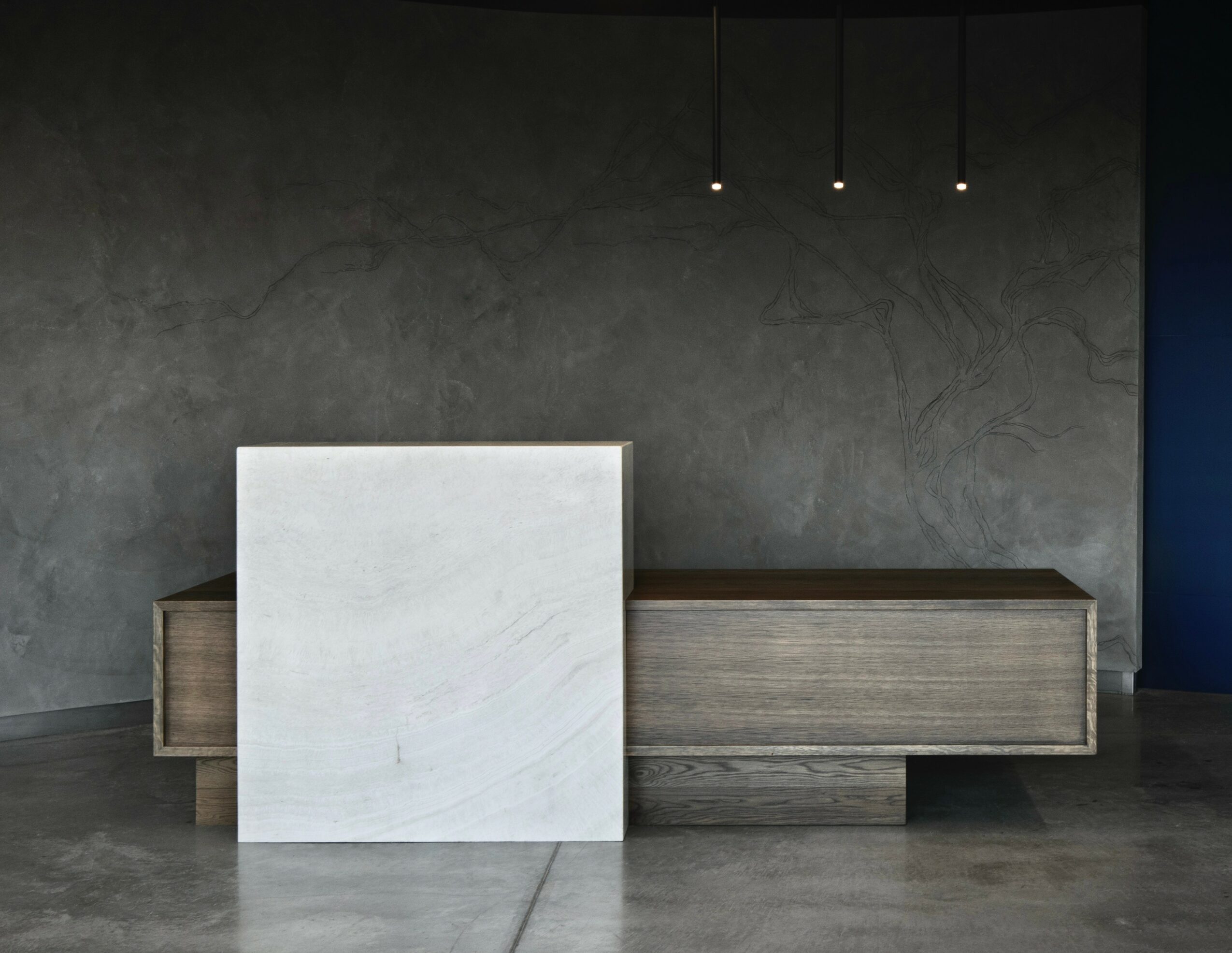 Modern reception desk with a white panel in front, against a dark textured wall and three hanging lights.
