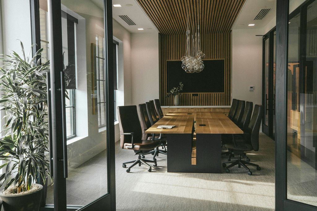 Modern conference room with a long wooden table, black chairs, large windows, and a chandelier, viewed through glass doors.