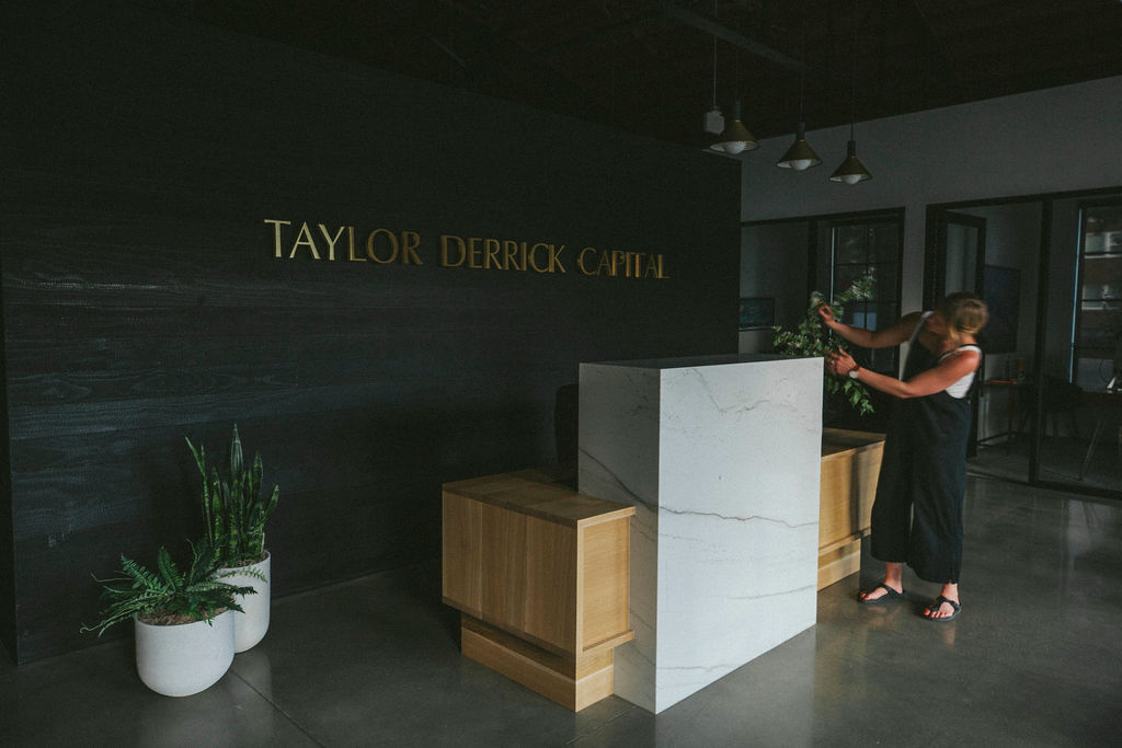 Two women take a selfie in front of a black wall with gold text reading 'TAYLOR DERRICK CAPITAL'.