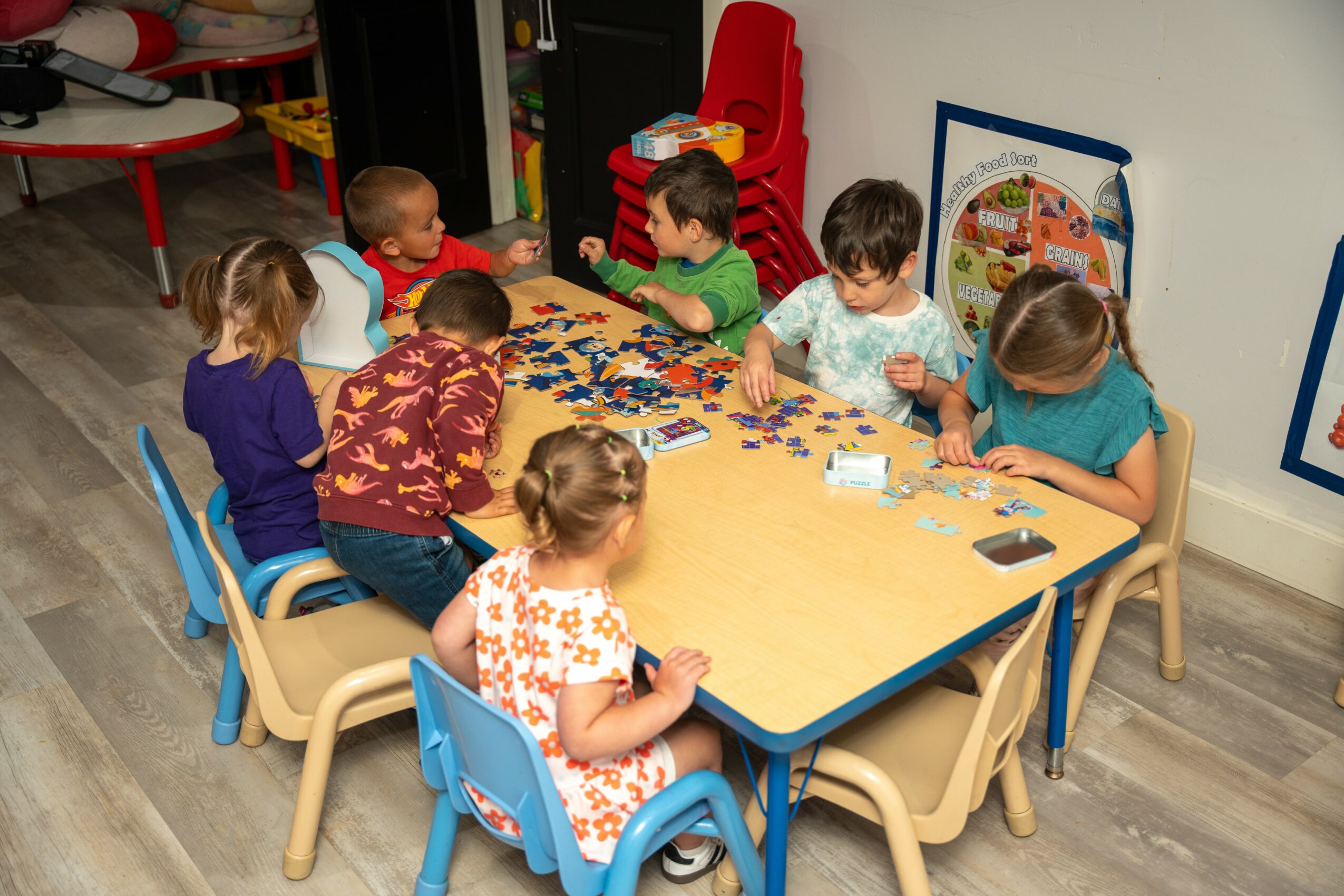 Children sit around a table playing with puzzle pieces in a classroom, with colorful chairs and educational posters on the wall.