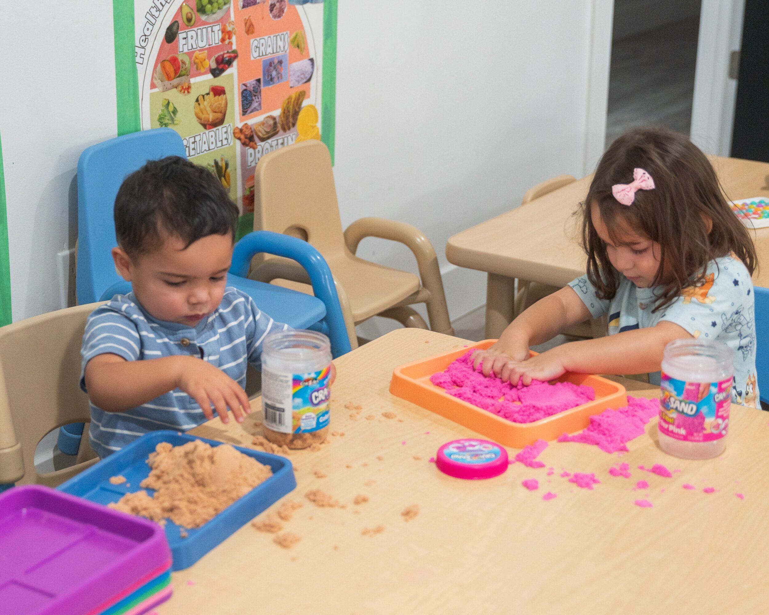 Two children playing with pink and beige sensory materials at a table, with containers and toys nearby.