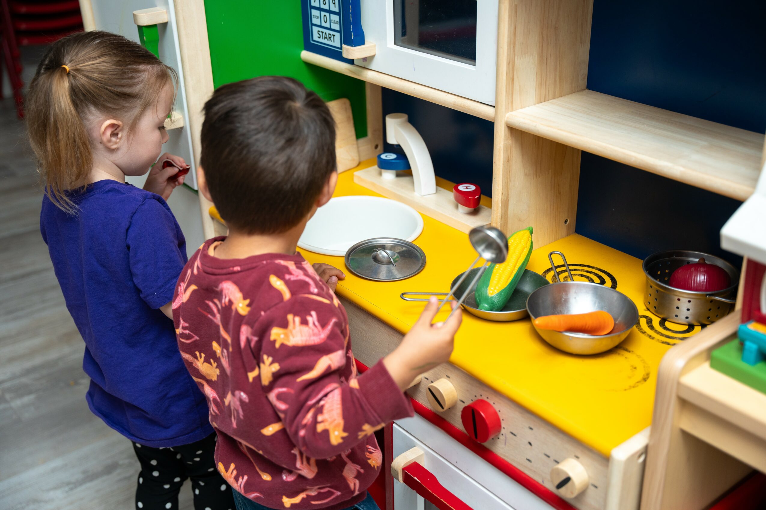 Two children stand at a toy kitchen set with a yellow countertop, playing with pots and utensils.