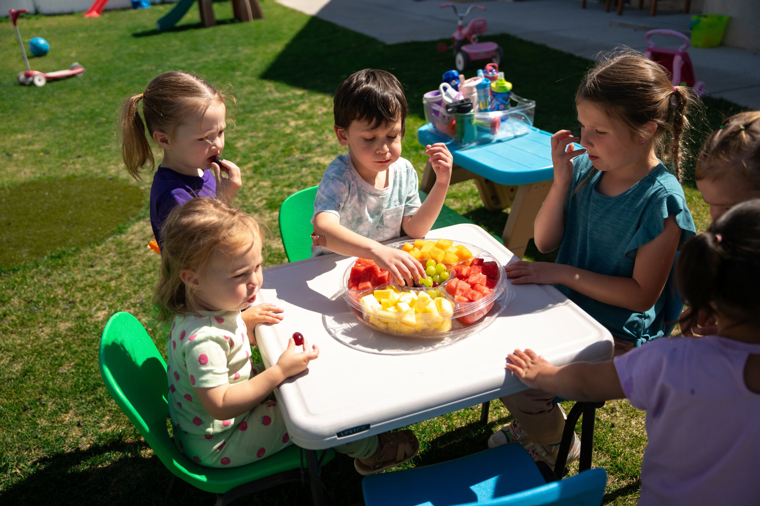 Children sit around a table outdoors, eating fruit and playing with toys, with grass and playground equipment in background.