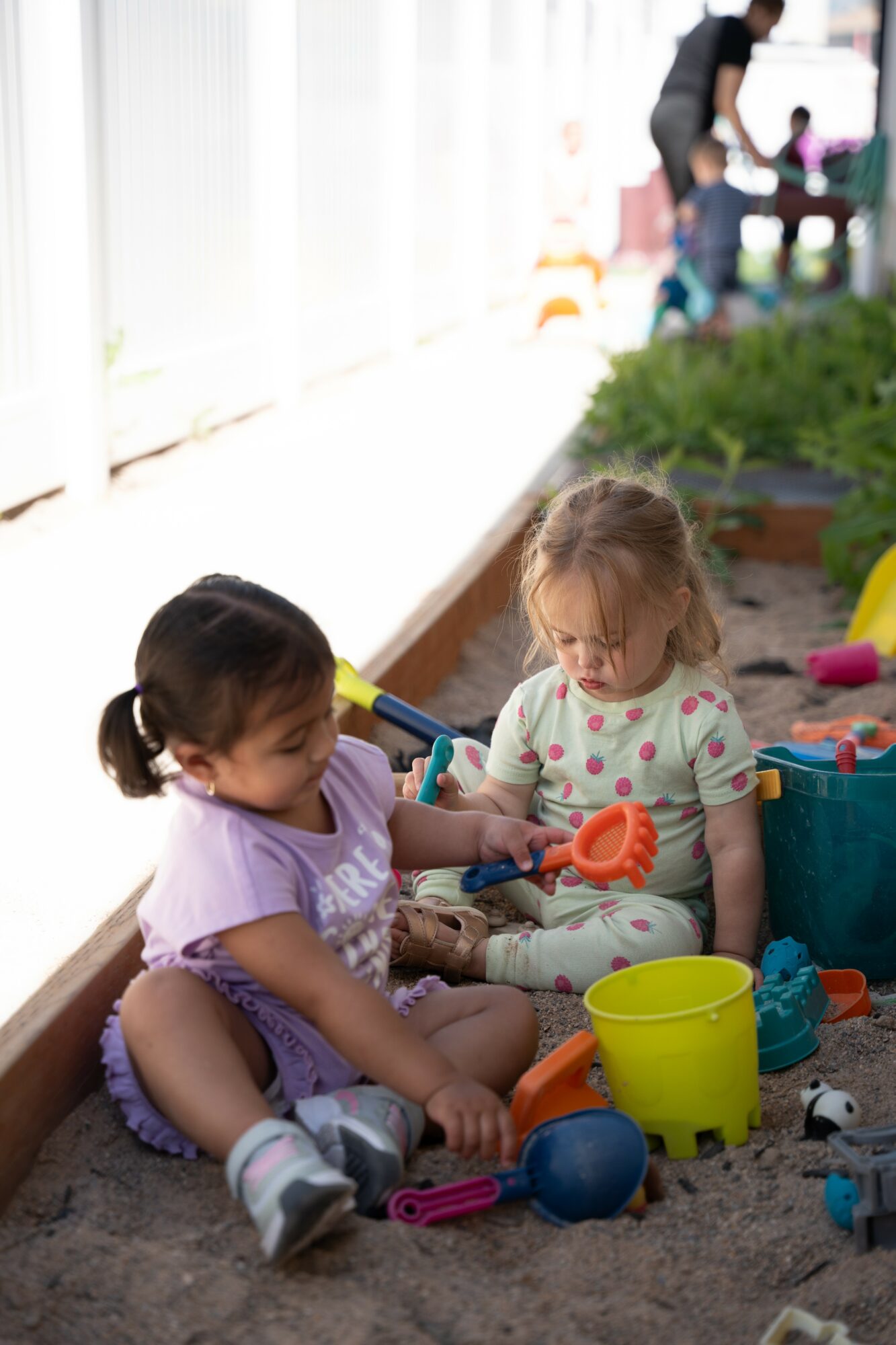 Two young girls playing with sand toys in a sandbox, surrounded by colorful buckets and toys, with people in the background.