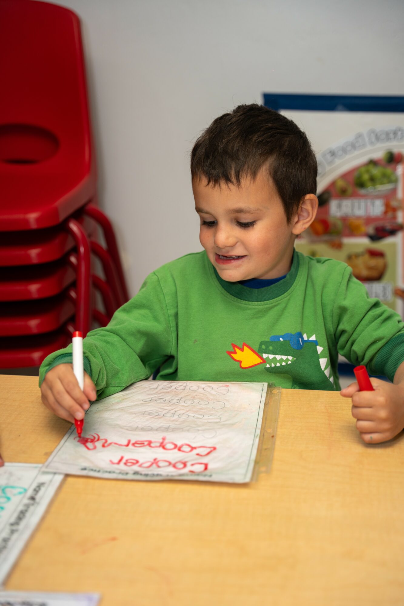 Young boy in green shirt writing in a notebook at a table, with red chairs stacked in the background.