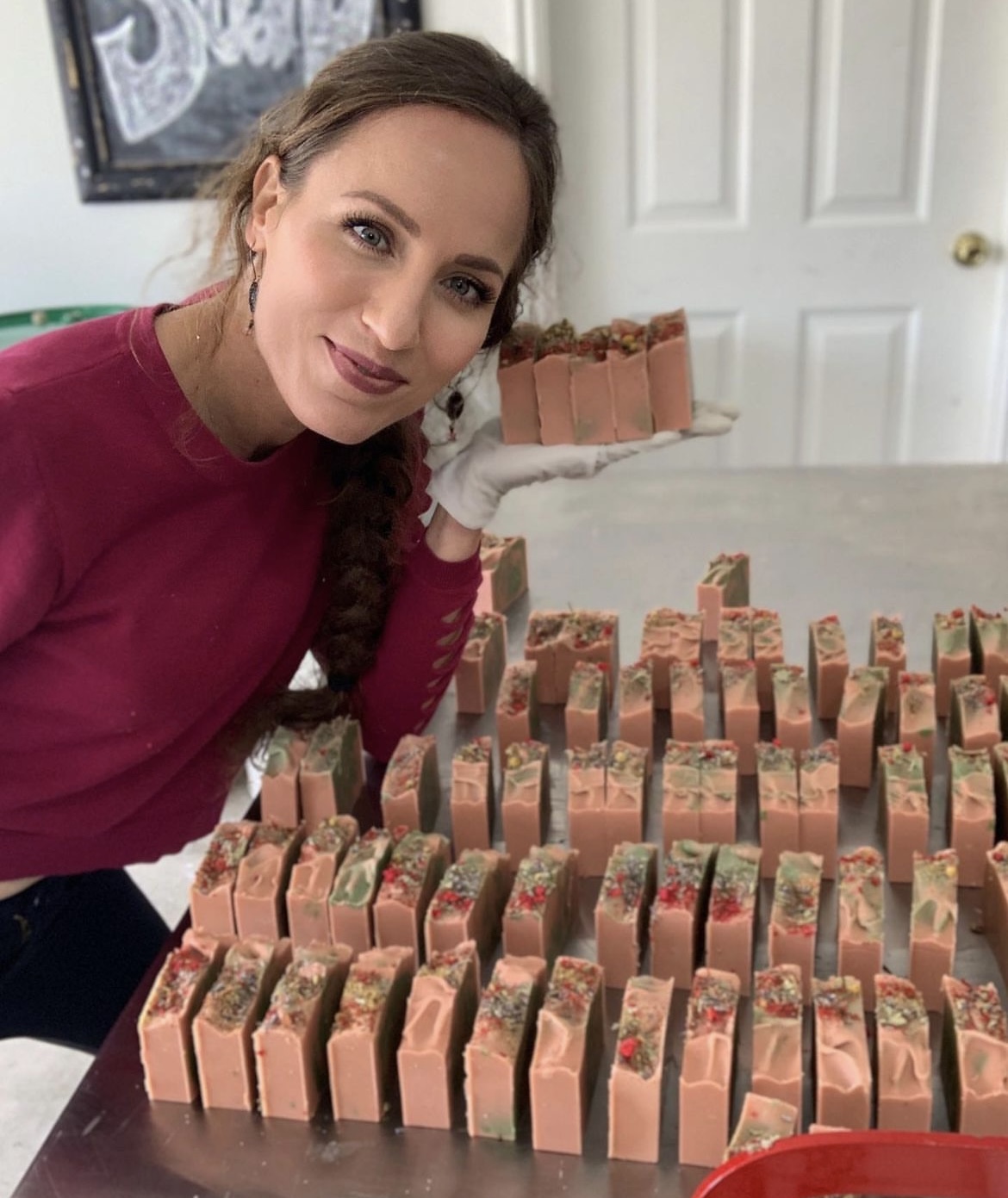 Woman with braided hair and pink sweater holding soap bar, surrounded by soap bars on table, in a room with white door.